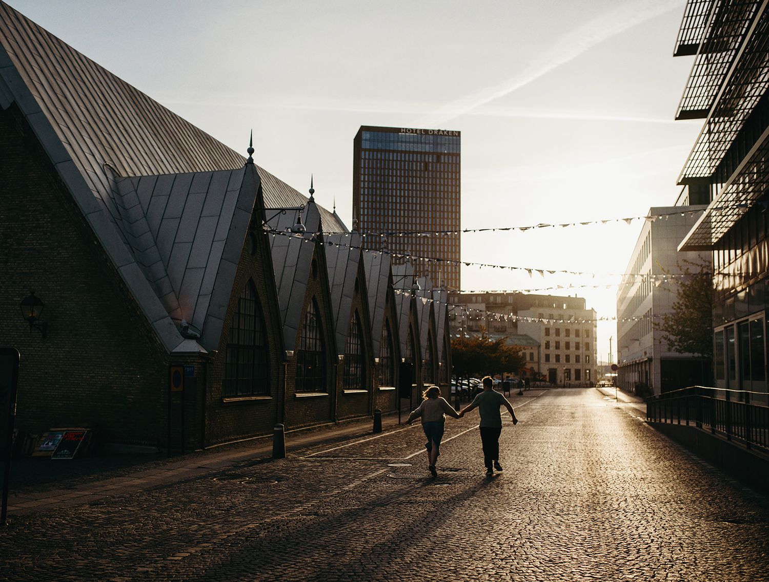 Two silhouetted figures walk down an urban street at sunset with modern buildings and string lights overhead.