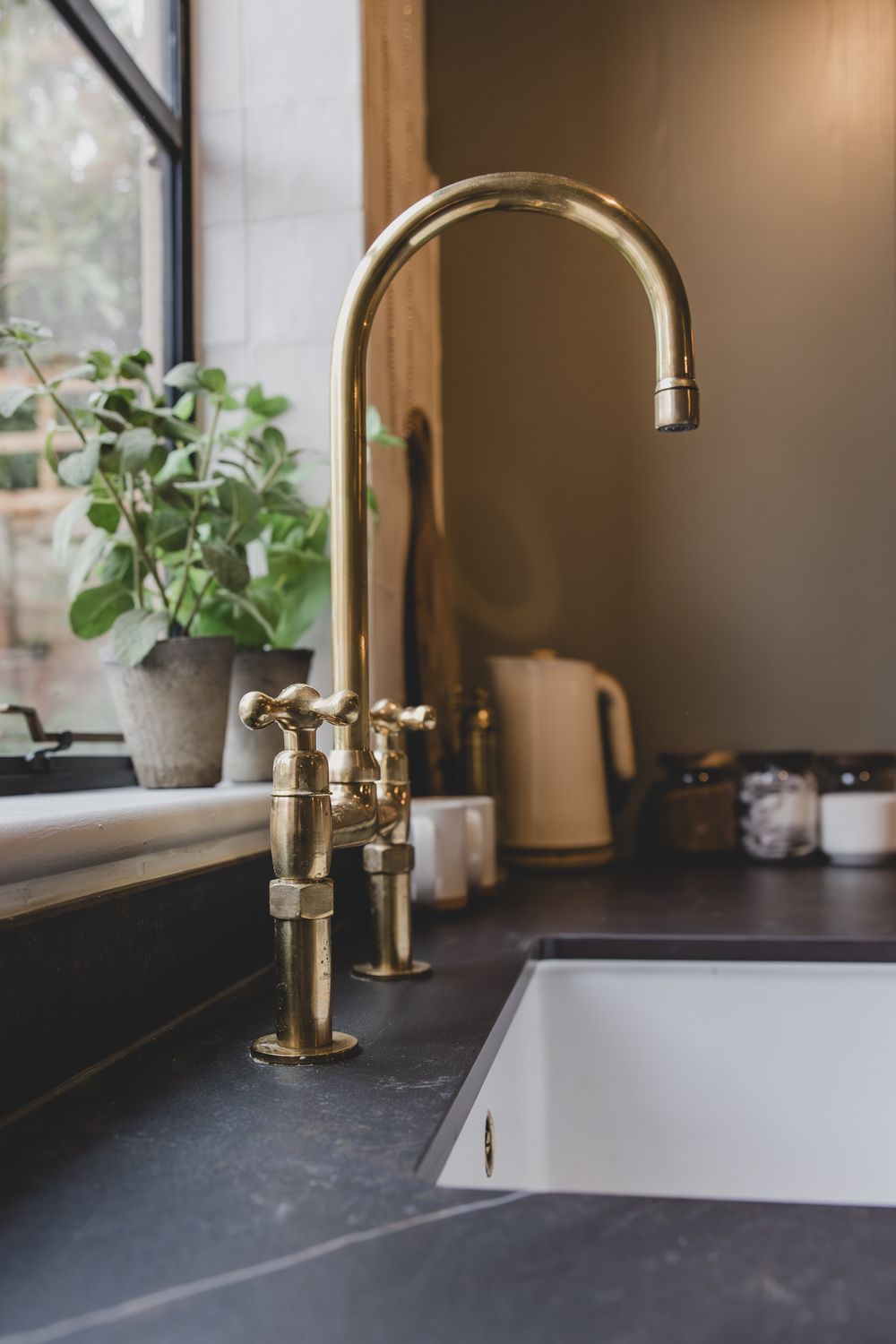Elegant brass bridge kitchen faucet with cross handles on dark slate countertop near window with green plants.