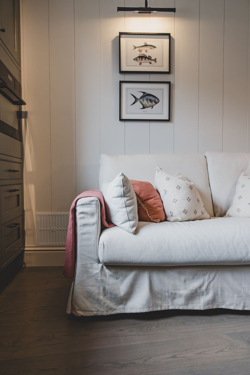 White slipcovered sofa with pink and patterned pillows beneath two framed fish prints on a shiplap wall.