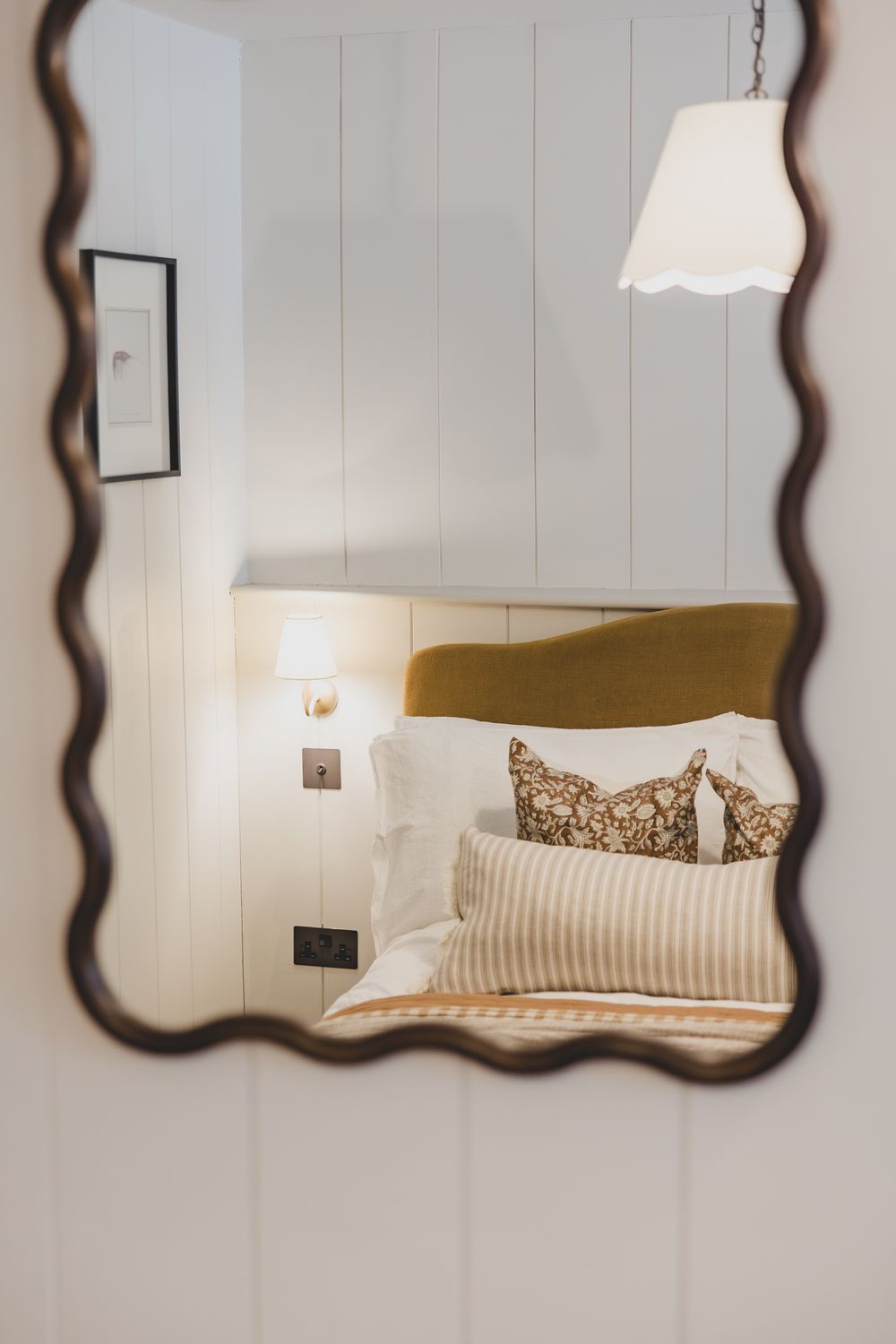 Wavy bronze mirror reflecting a cozy bedroom with mustard headboard, striped pillows, and warm wall sconce lighting.