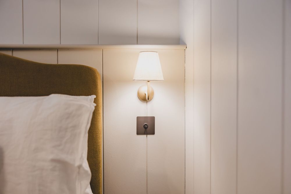 A cozy bedroom corner with a glowing wall sconce lamp beside an upholstered headboard with white pillows.