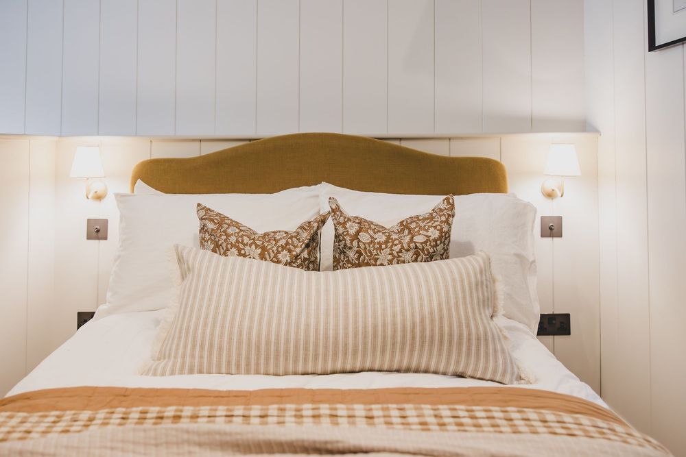 Elegant bedroom with gold velvet headboard, striped lumbar pillow, floral cushions, and white bedding with warm accent lighting.