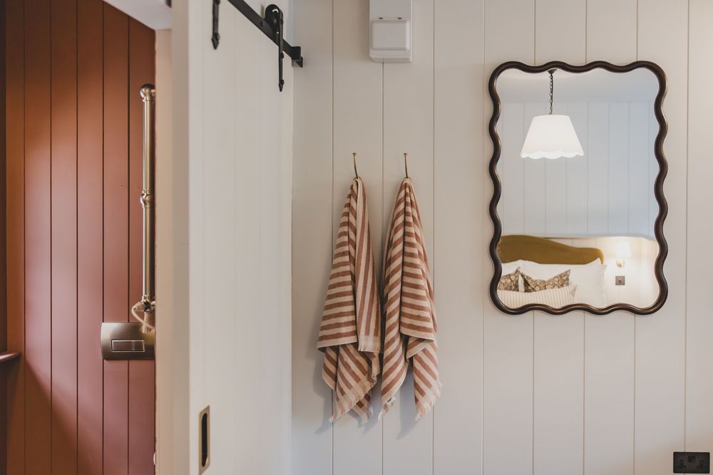 Striped towels hang on hooks beside a wavy mirror in a cozy white shiplap bathroom with a barn door.