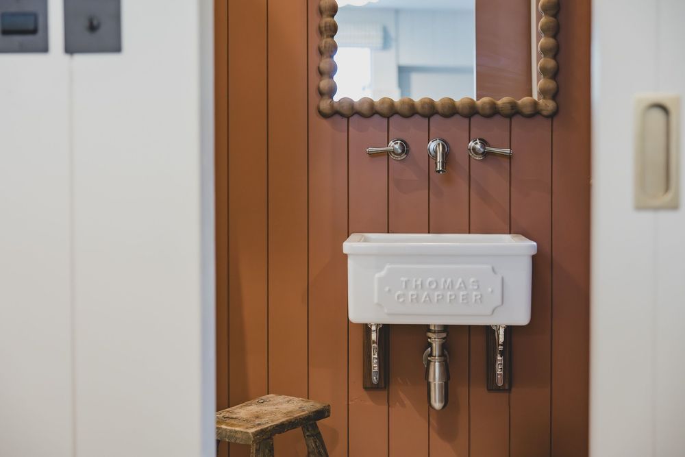 Thomas Crapper white wall-mounted sink with chrome faucets on wood-paneled wall with beaded mirror above.