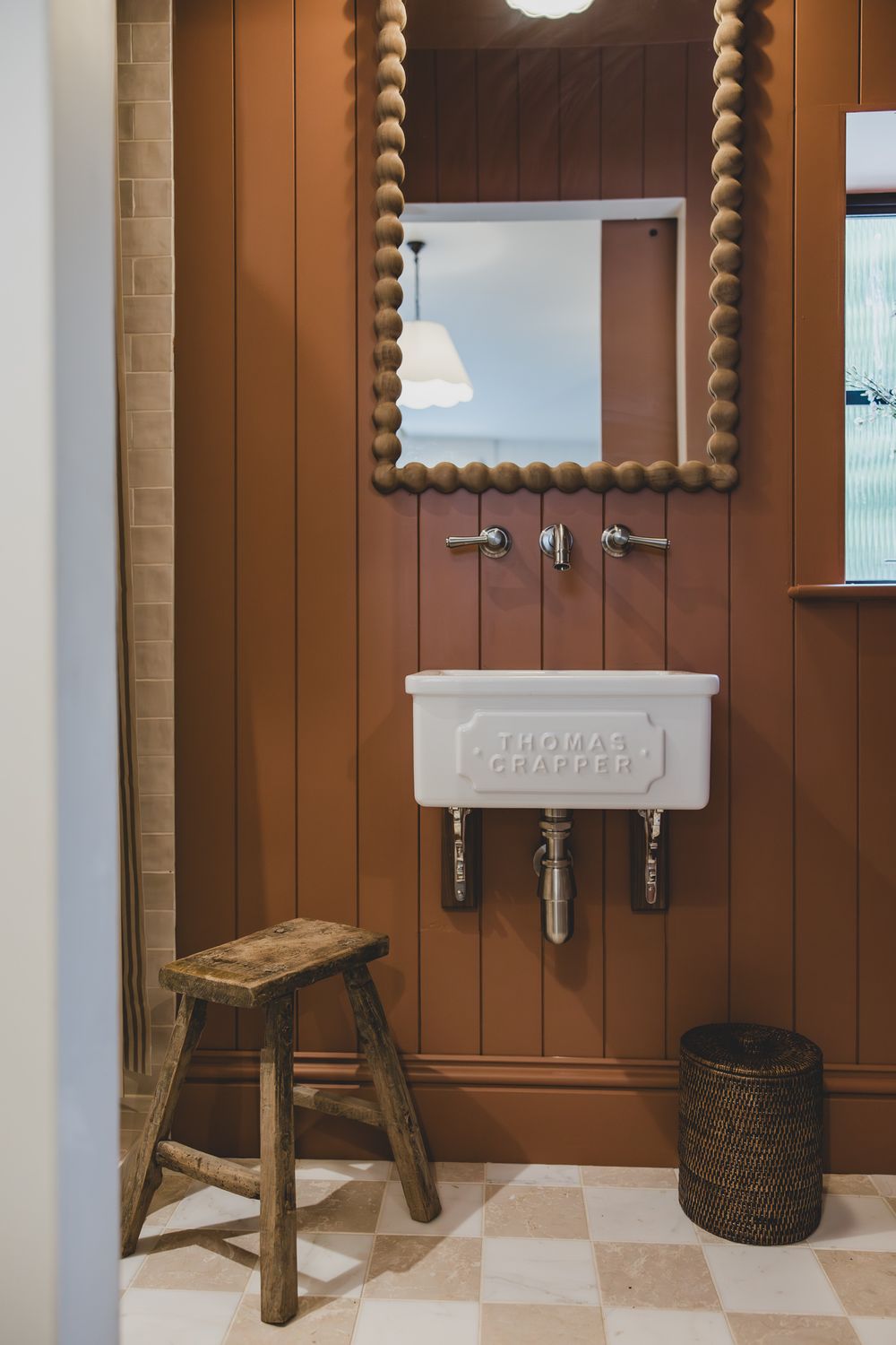 Rustic bathroom with brown wood paneling, Thomas Crapper white sink, beaded mirror, wooden stool, and checkered tile floor.
