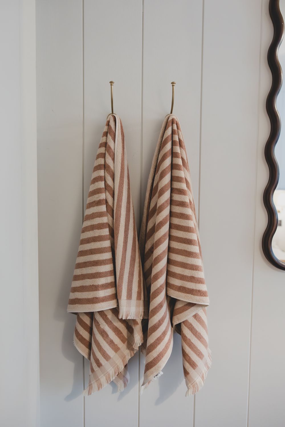 Two striped brown and white Turkish towels hanging on brass hooks on a white shiplap wall.