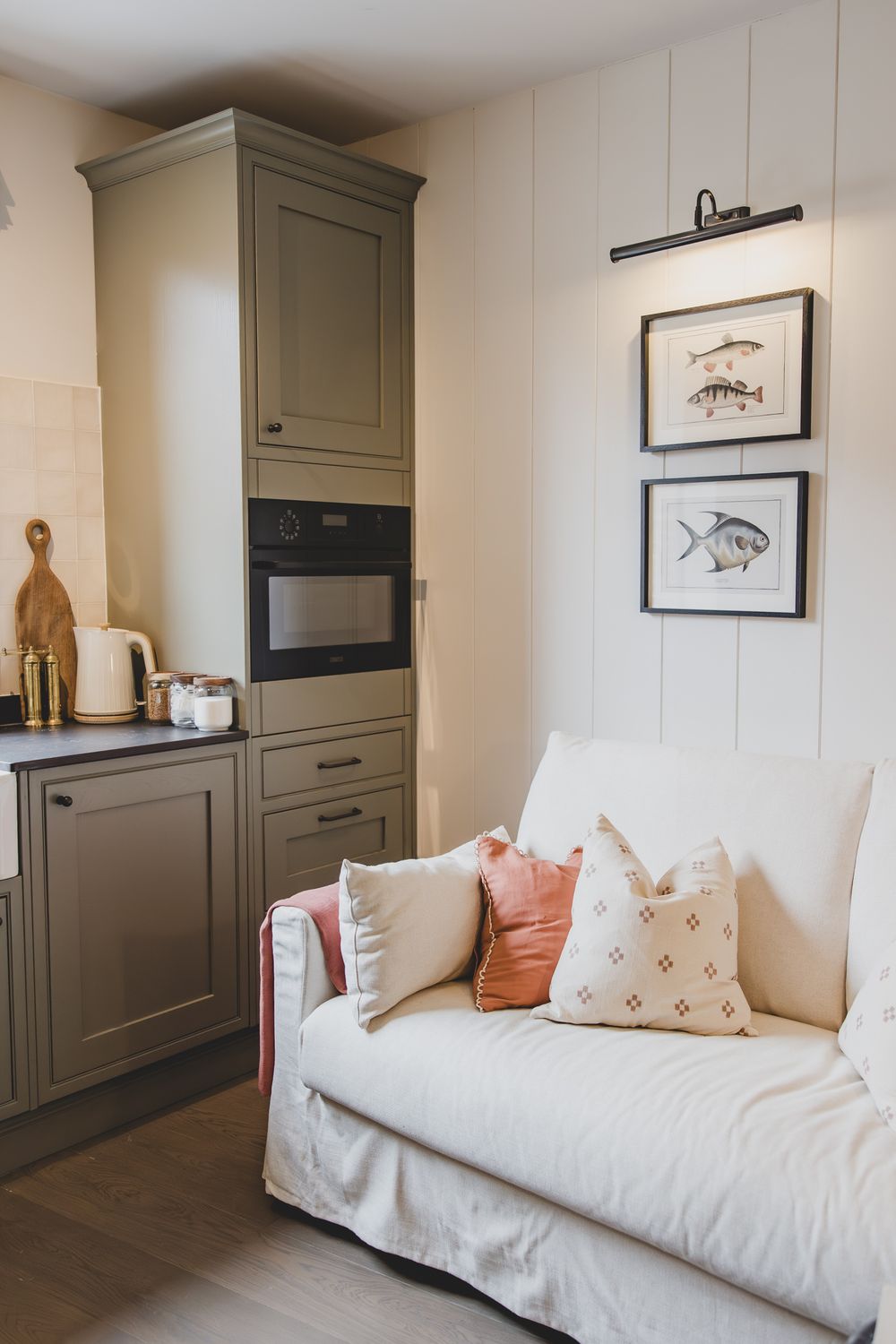 Cozy kitchen nook with olive green cabinets, built-in oven, white sofa with pink pillows, and framed fish prints on shiplap wall.