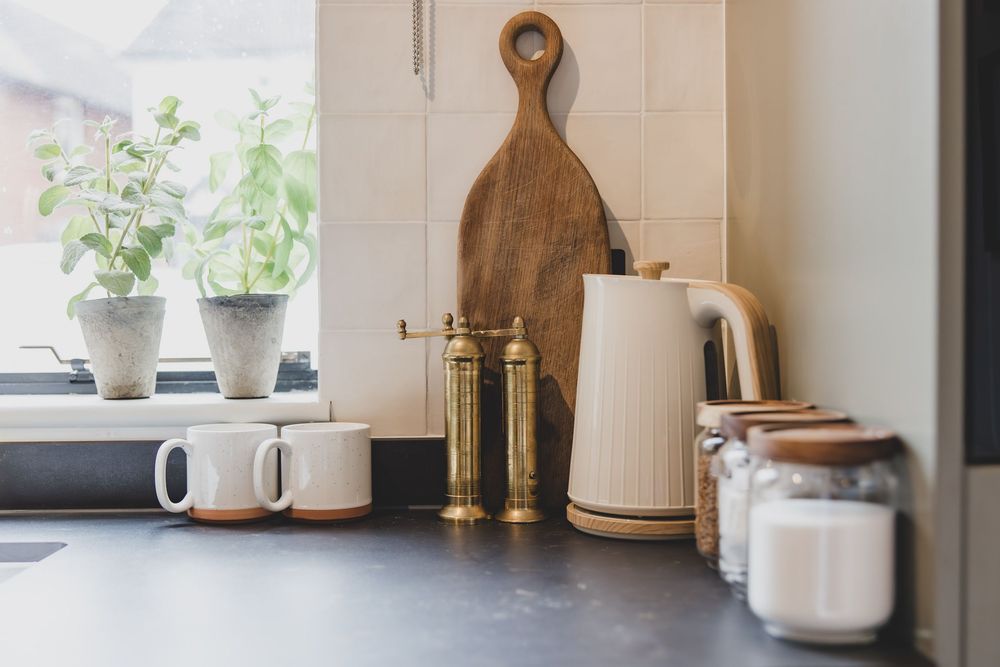 Cozy kitchen counter with brass pepper mills, wooden cutting board, white kettle, mugs, and potted plants by a window.