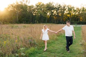 A couple holds hands while walking through a golden meadow at sunset with trees in the background.