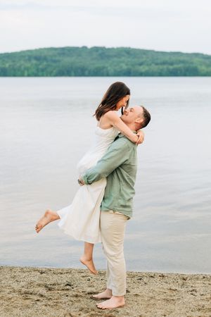 A couple embraces on a sandy beach with a scenic lake and forested hill in the background.