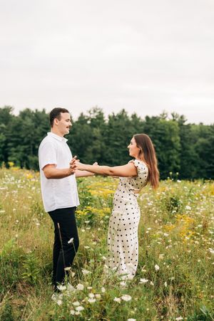 A couple stand together in a wildflower meadow with trees in the background during summer.