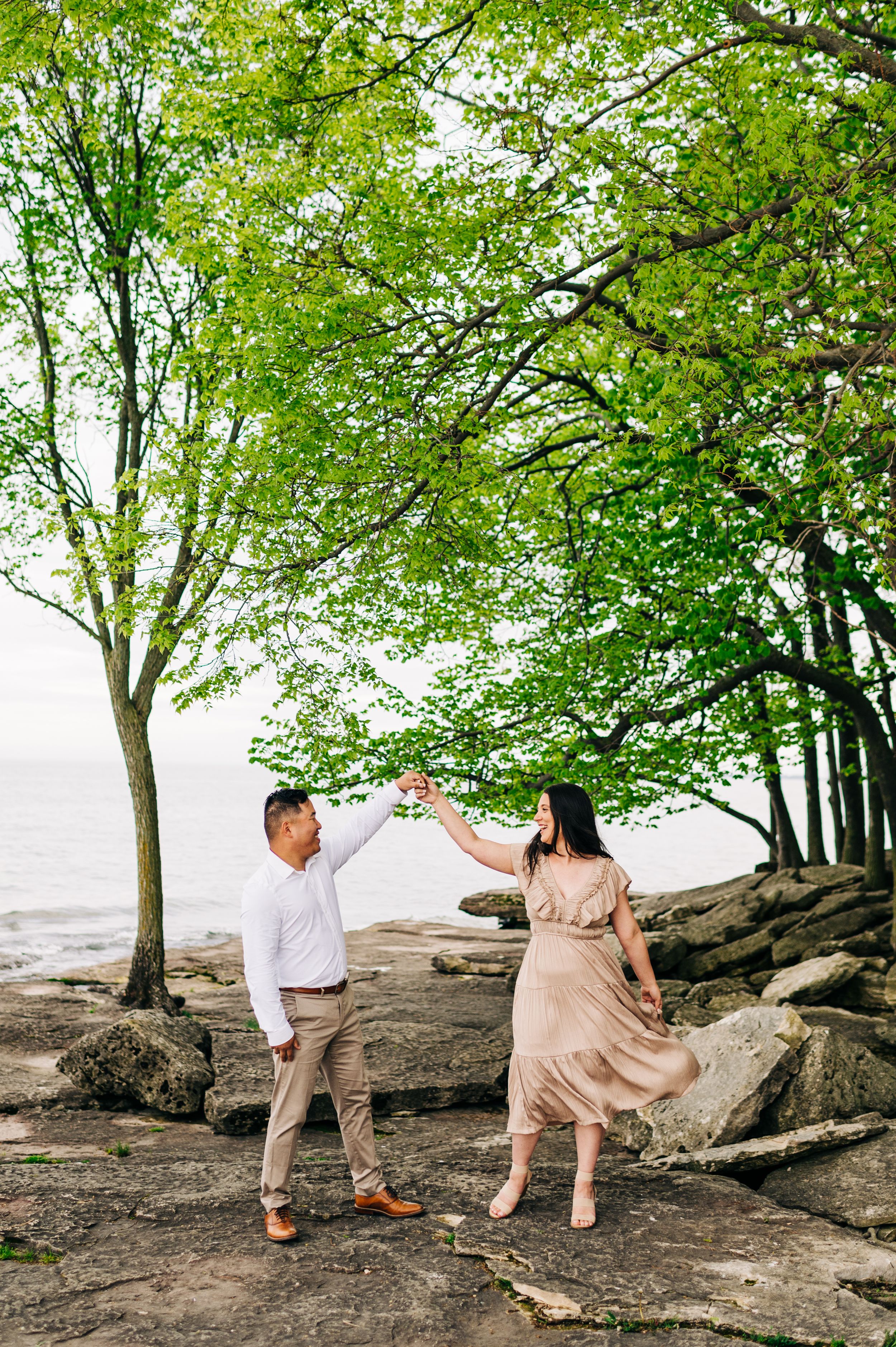 A couple dances together on rocky coastal shoreline under green leafy trees by the water on a bright day.