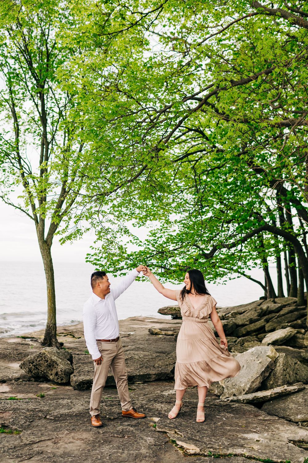 A couple dances together on rocky coastal shoreline under green leafy trees by the water on a bright day.