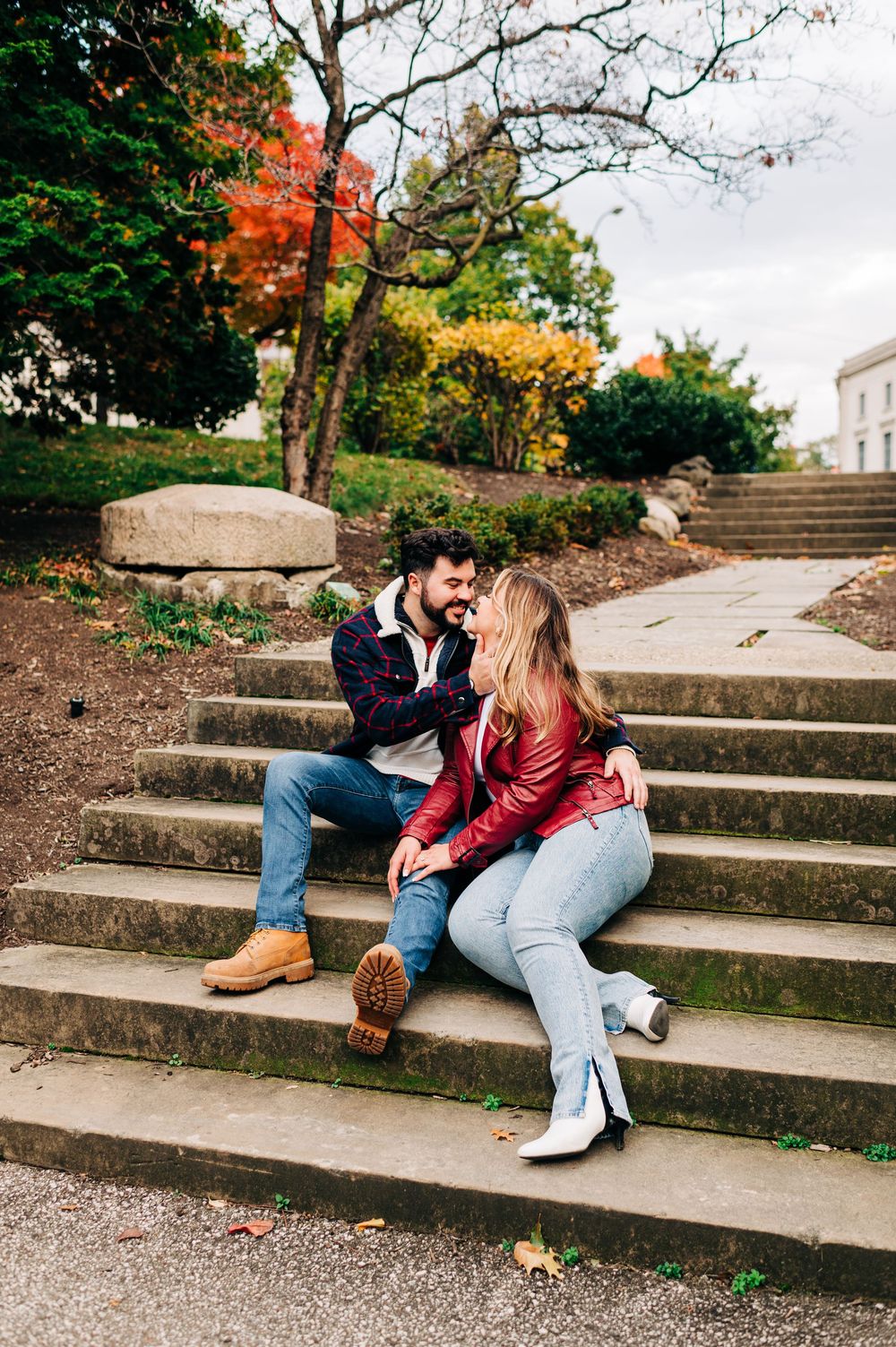 A couple sits together on concrete stairs during autumn with colorful fall foliage in the background.