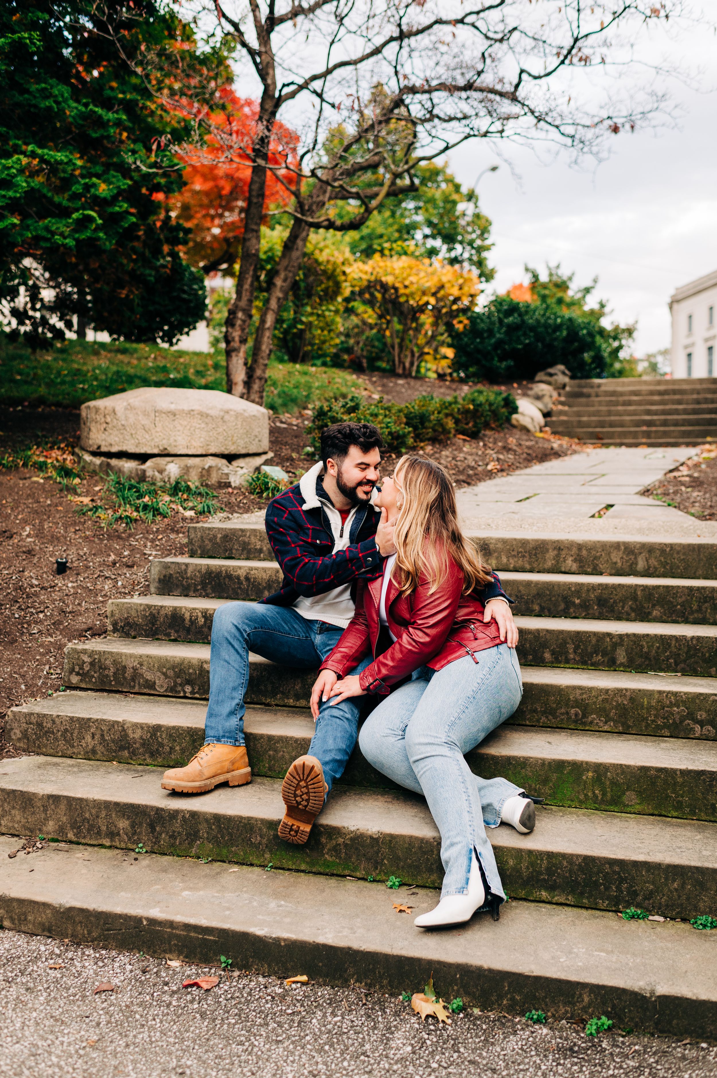 A couple sits together on concrete stairs during autumn with colorful fall foliage in the background.
