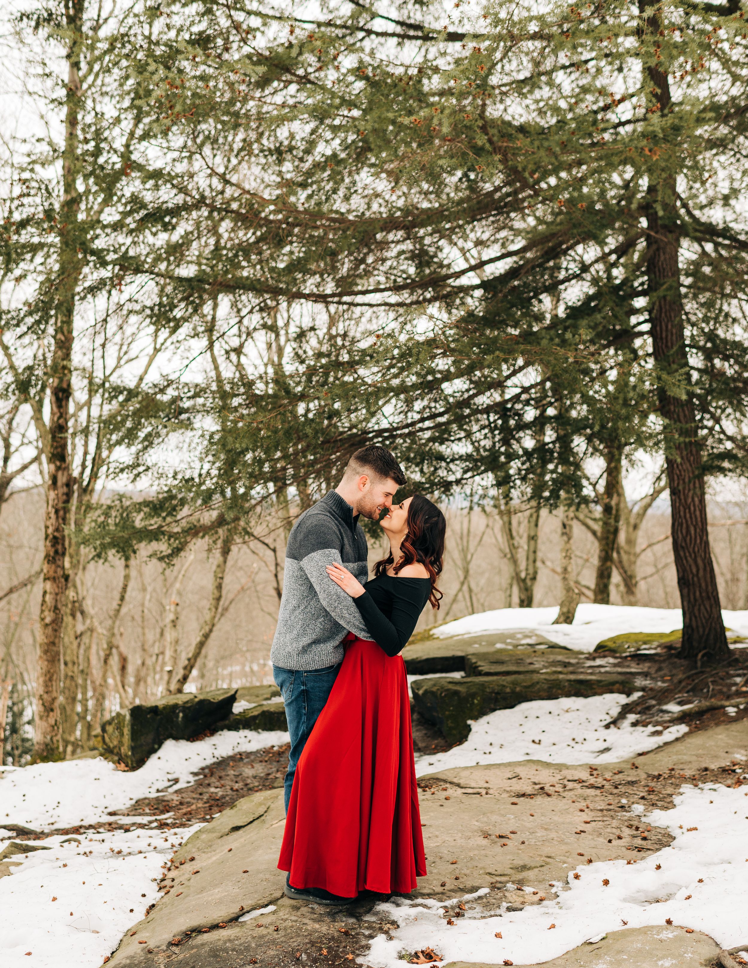 A romantic winter scene with a couple embracing on a snowy path surrounded by evergreen trees.