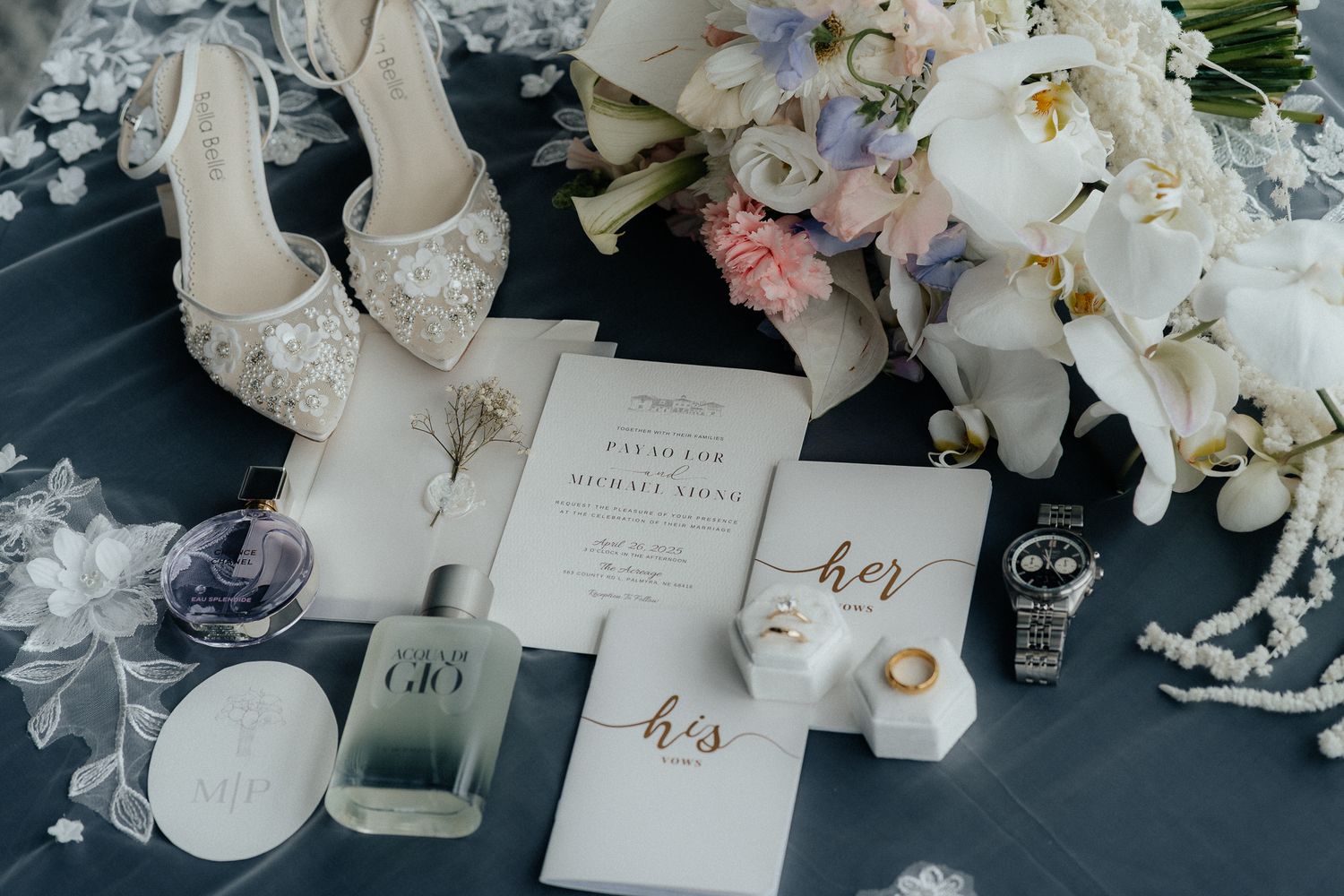 Wedding details flatlay with invitation, rings, shoes, and white floral bouquet on dark surface.