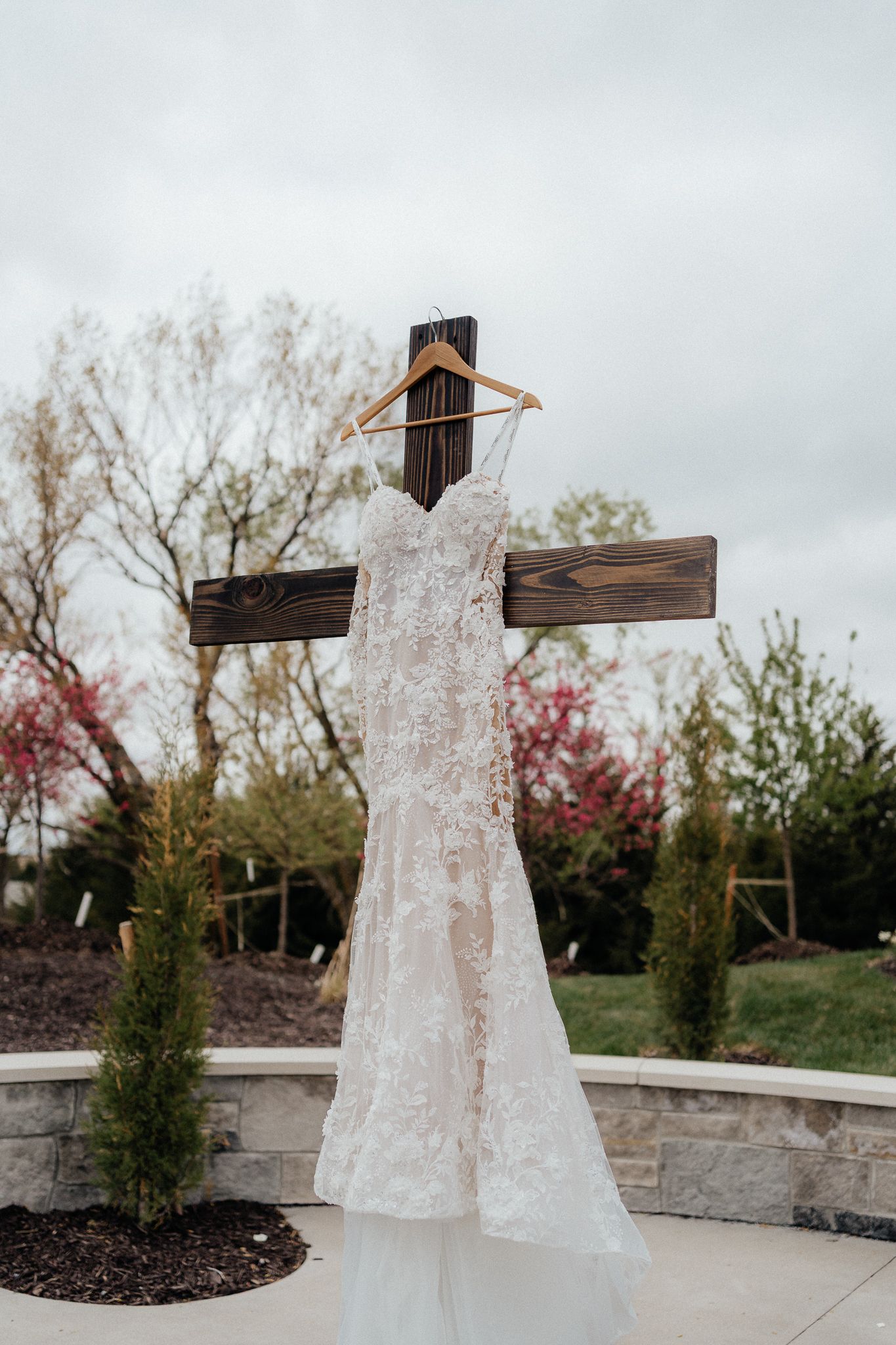 An elegant lace wedding dress displayed on a wooden cross-shaped hanger outdoors with autumn foliage in the background.