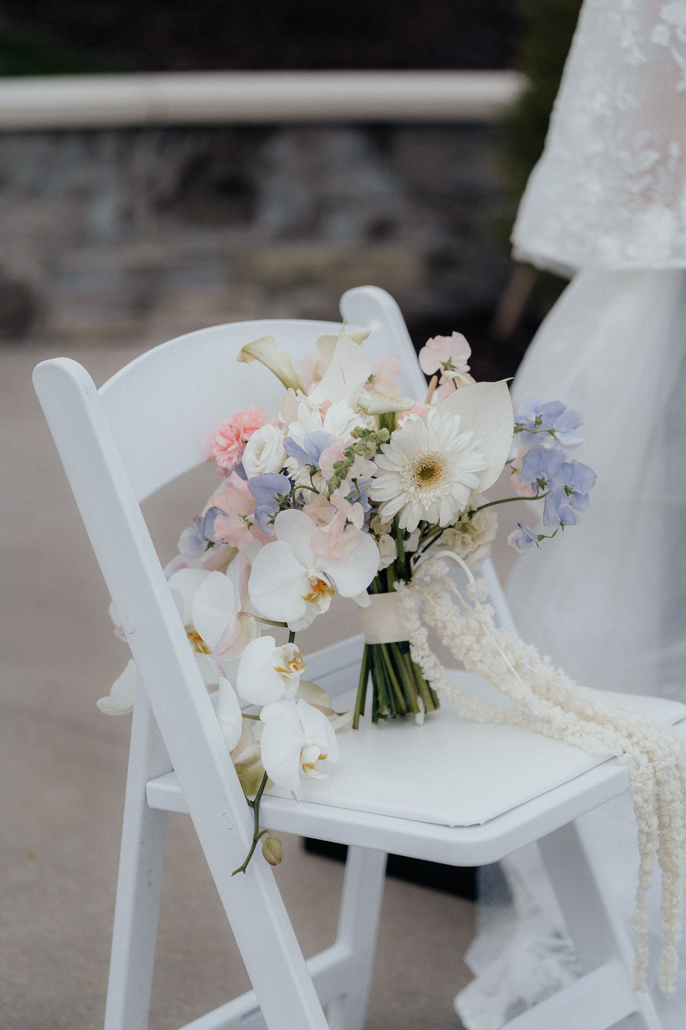 A delicate pastel wedding bouquet with white orchids and pink flowers rests on a white folding chair.