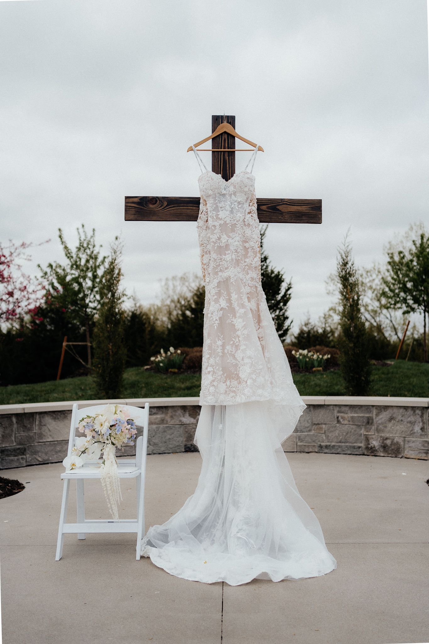 A white lace wedding dress hangs elegantly on a wooden cross against an overcast sky with trees in the background.