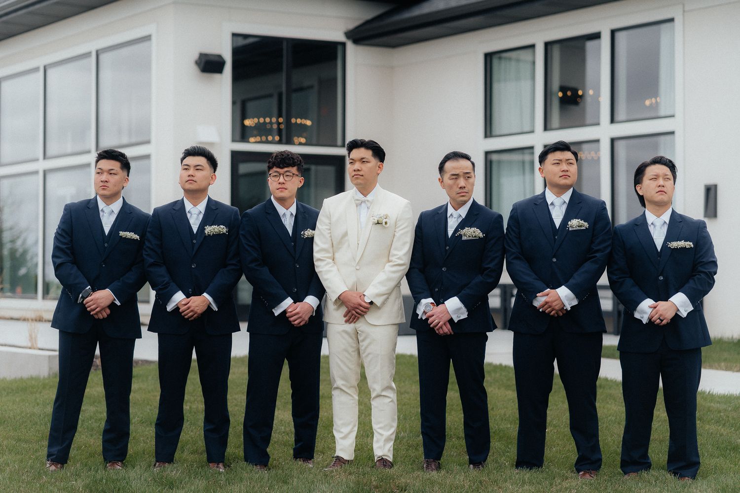 A wedding party in matching navy suits stands in a row outdoors with one member wearing cream colored attire.