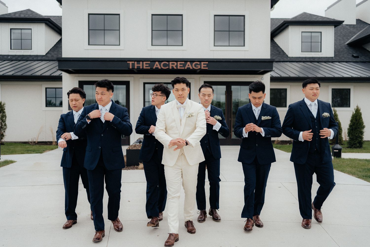 Wedding party in navy suits and light colored suit poses together outside The Acreage venue building.