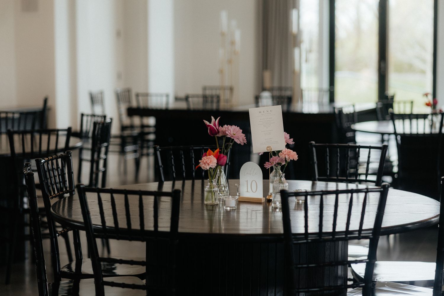 A reception venue set with round tables, black chiavari chairs, and small floral centerpieces in a bright room.