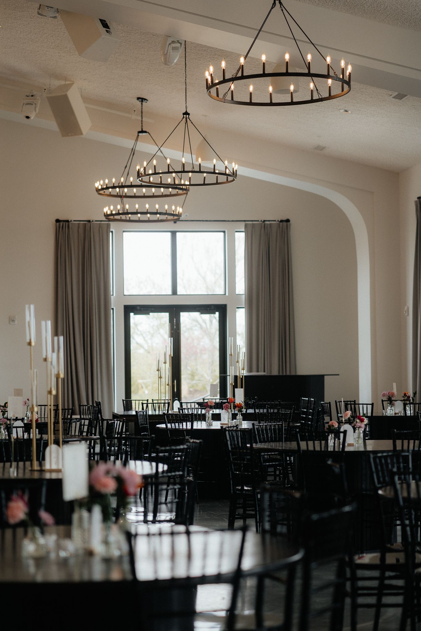 Elegant wedding venue dining room with large circular chandeliers, tall windows, and dark wood chairs in an upscale setting.