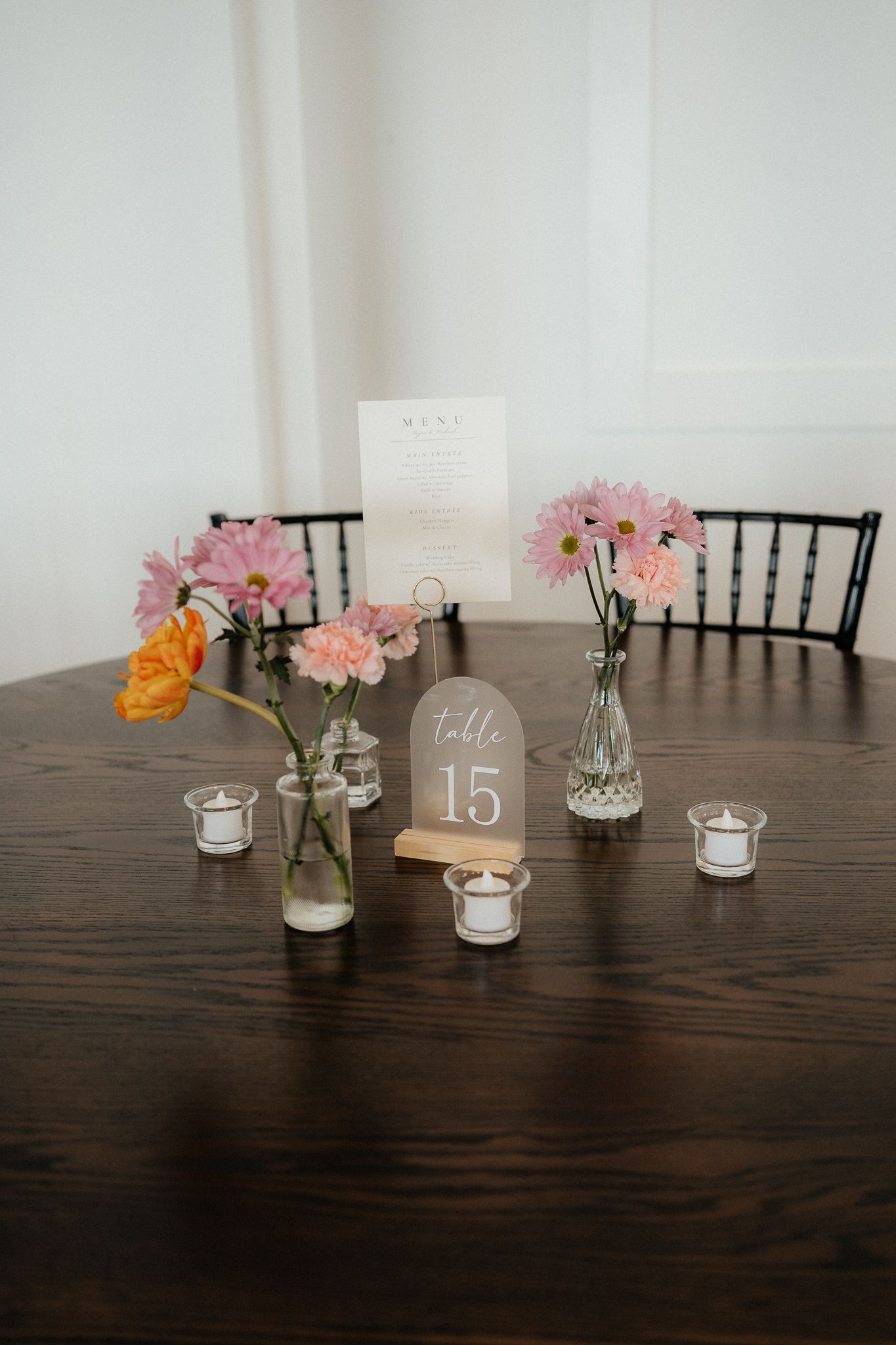 A rustic wedding table setting with pink flowers in vases, candles, and a table number 15 on a dark wooden surface.