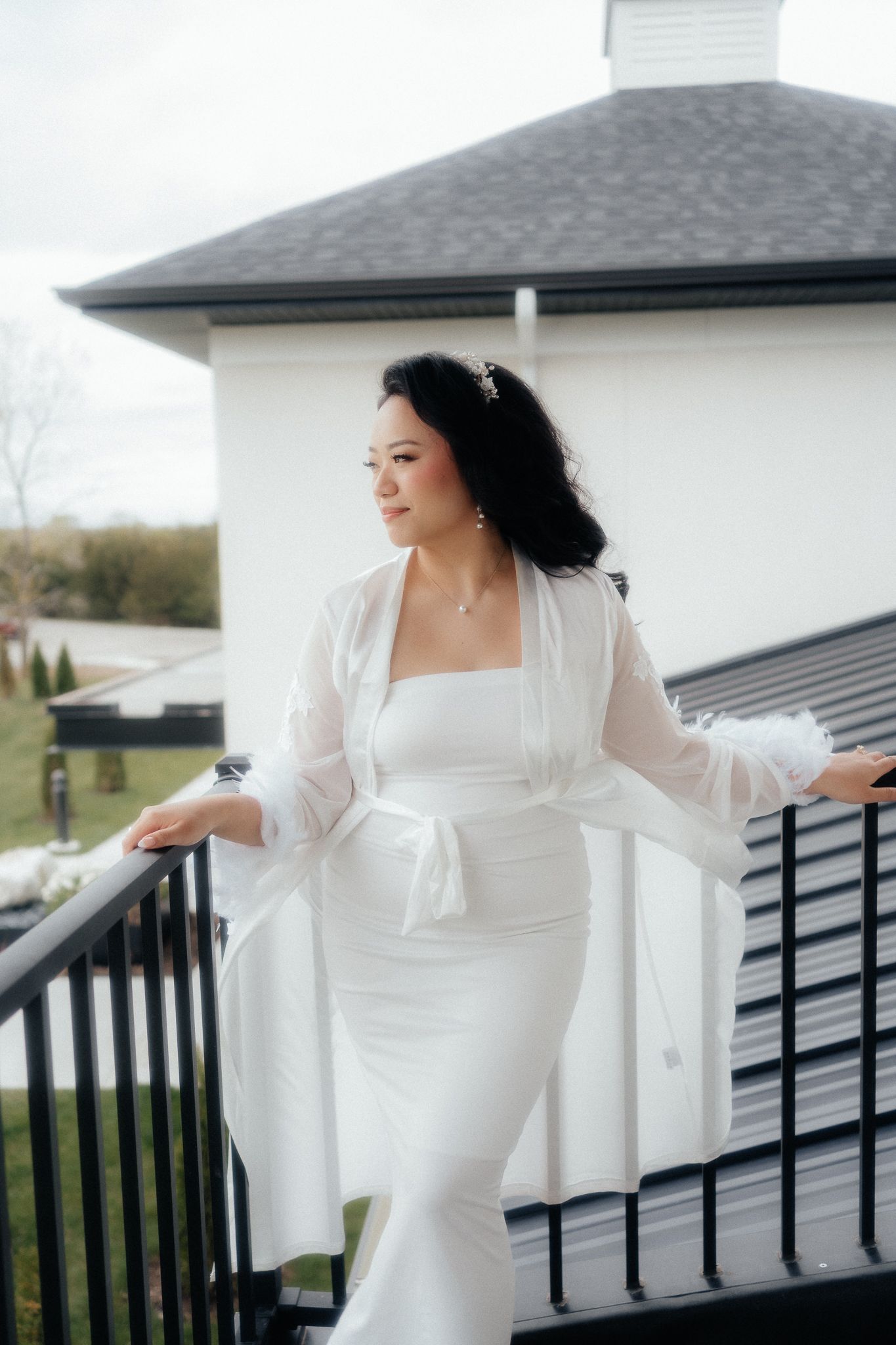 A stunning bride in a white wedding dress and sheer robe poses elegantly on a balcony of a white building.