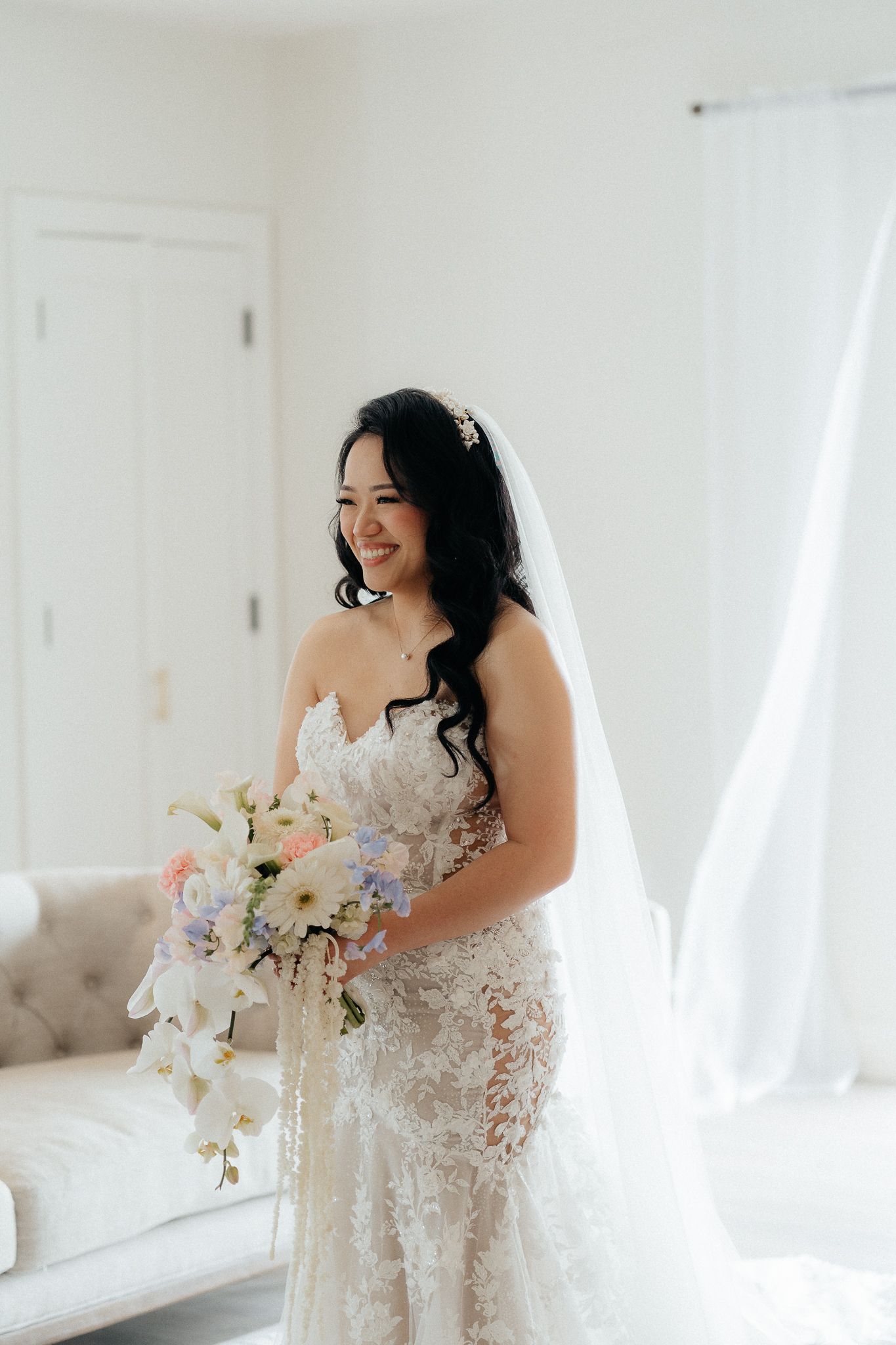 Bride in an elegant lace wedding dress holds a light pink and white bouquet while smiling in a bright room.