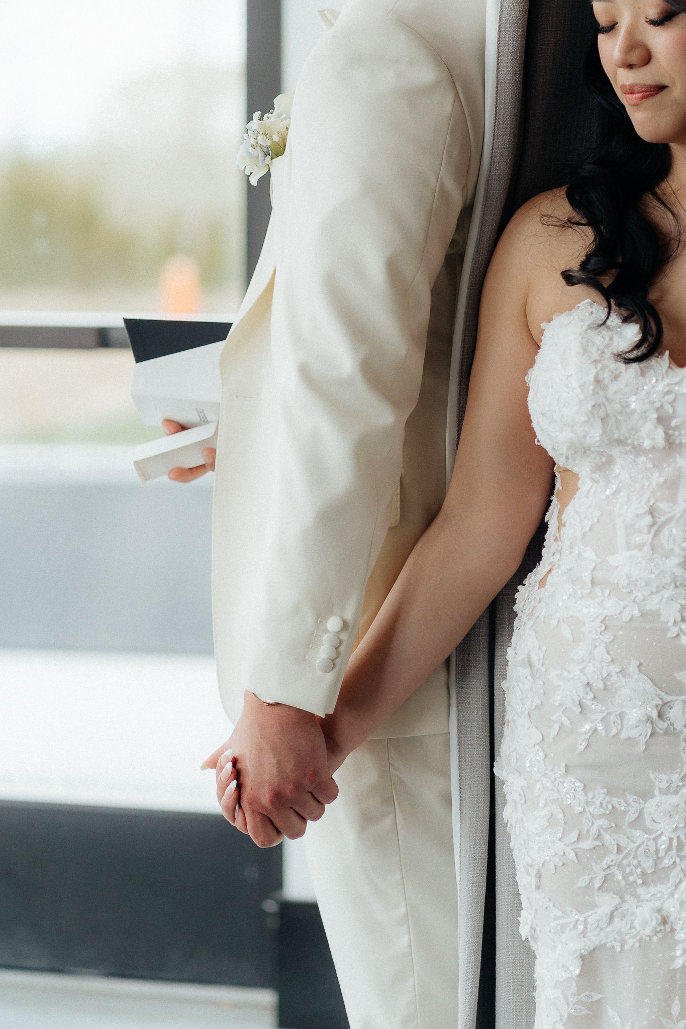 Close-up of wedding couple holding hands, with bride in lace dress and groom in white suit standing by window.