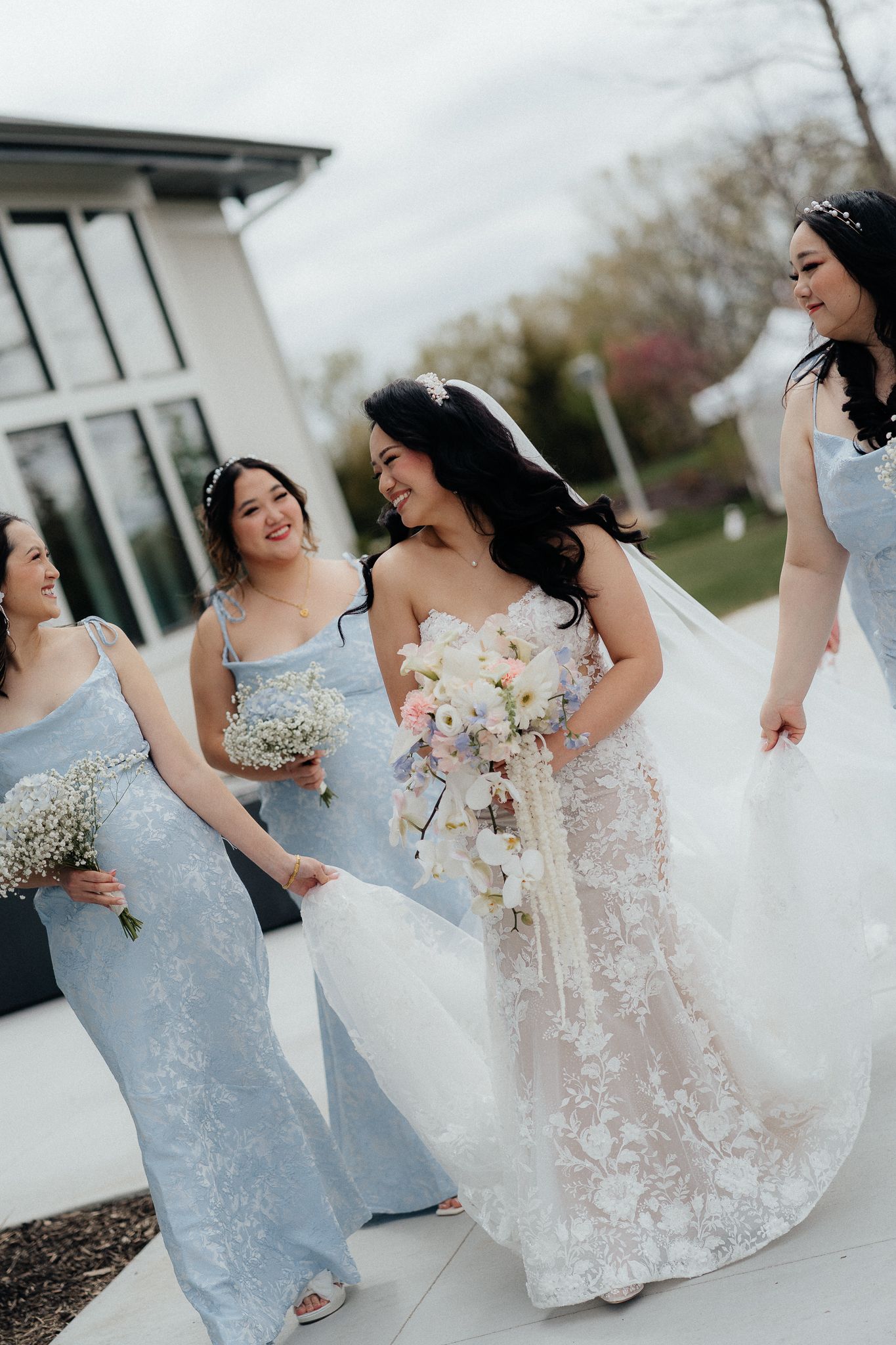 A bridal party in light blue bridesmaid dresses and a lace wedding gown share a candid moment outside.