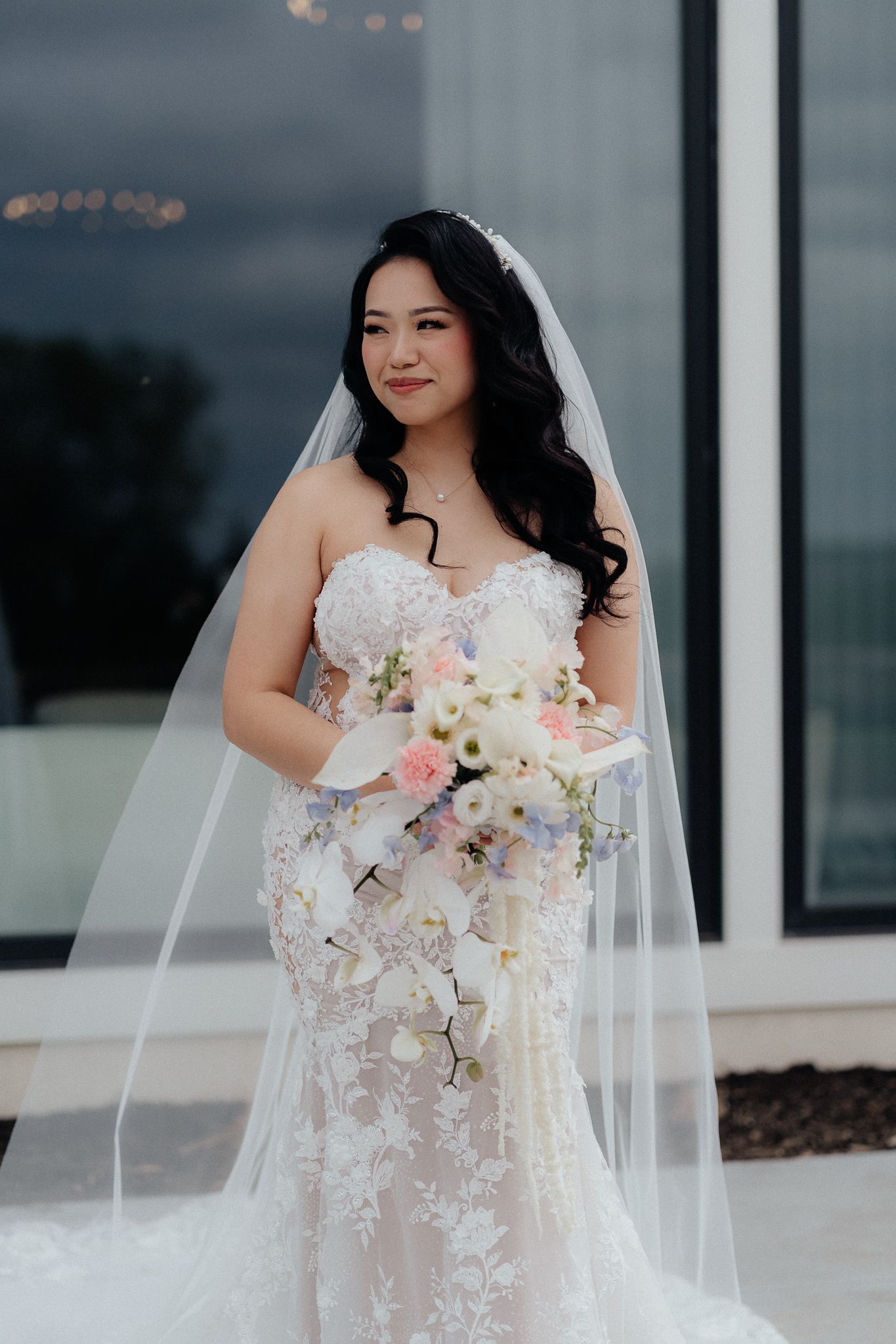 A bride in a strapless lace mermaid gown holds a delicate pastel bouquet against a stormy sky.