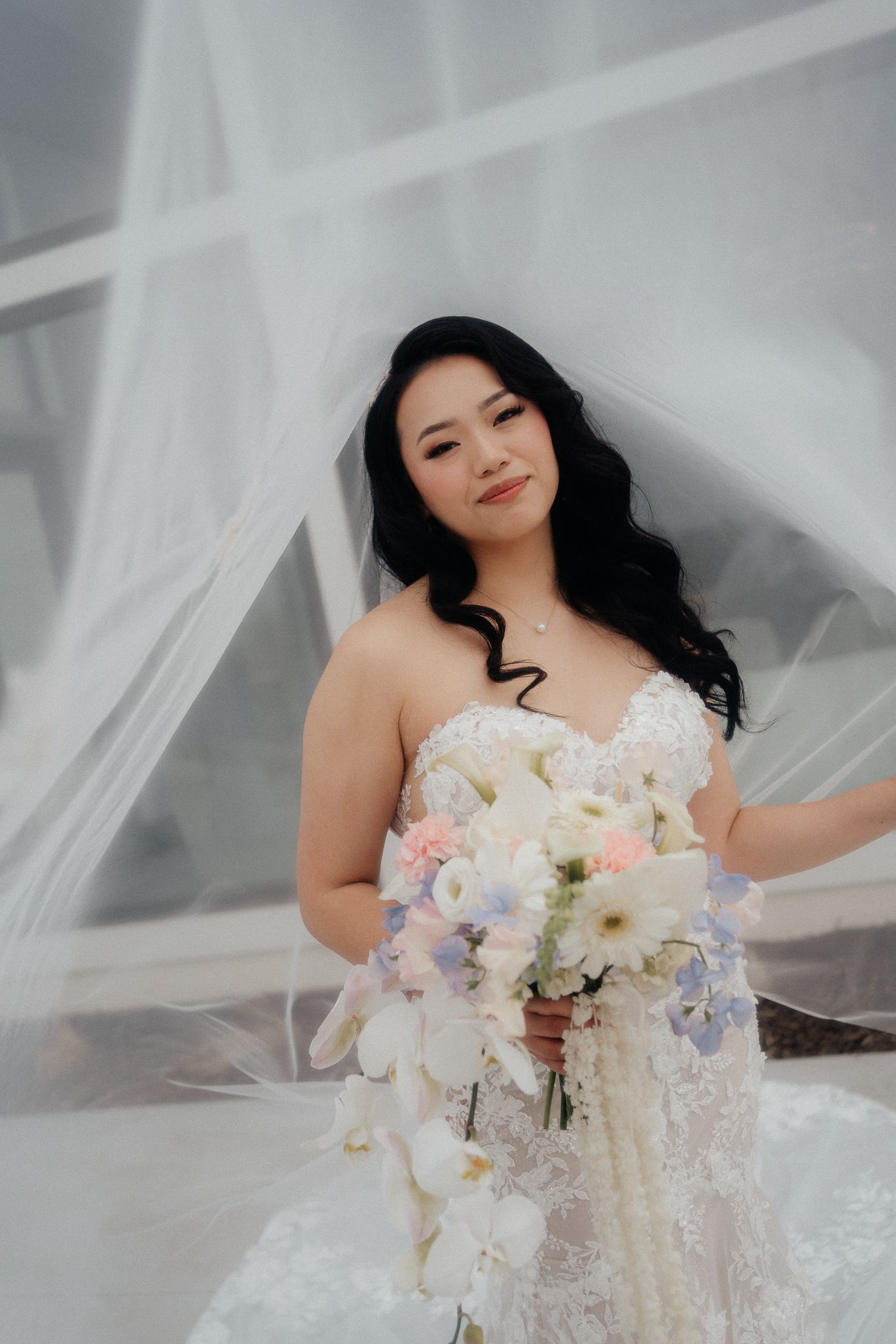 A bride in a lace wedding dress holds a bouquet of white orchids and pastel flowers under a flowing veil.