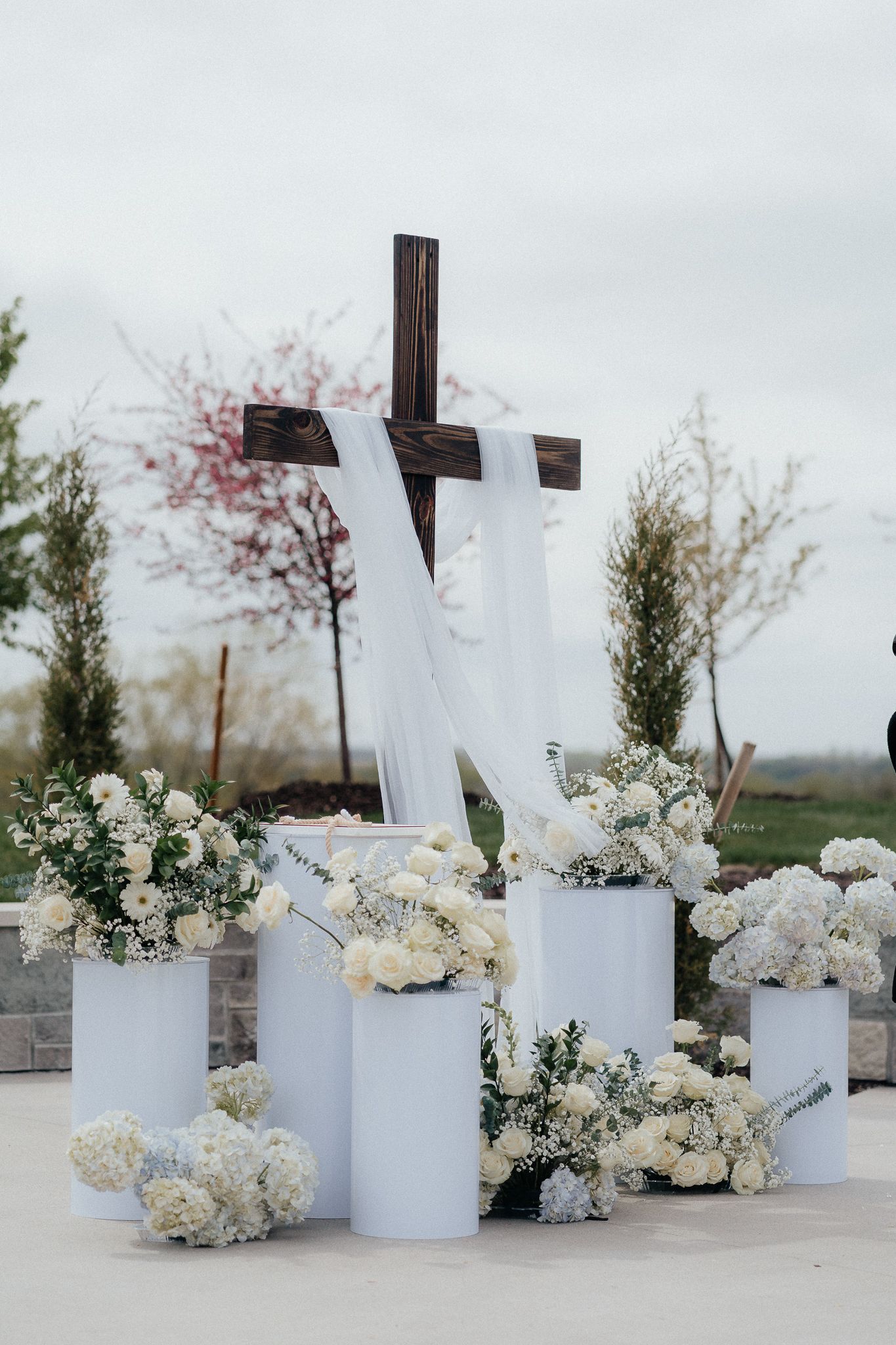 White floral arrangements with cross at outdoor wedding ceremony setup.