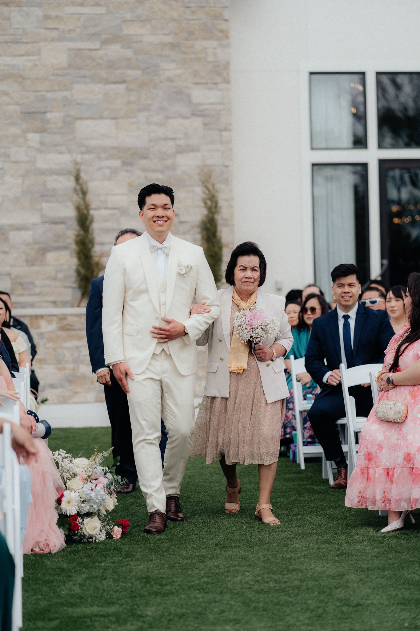Wedding guests watch as a couple walks down the aisle on a grassy lawn with a stone wall backdrop.