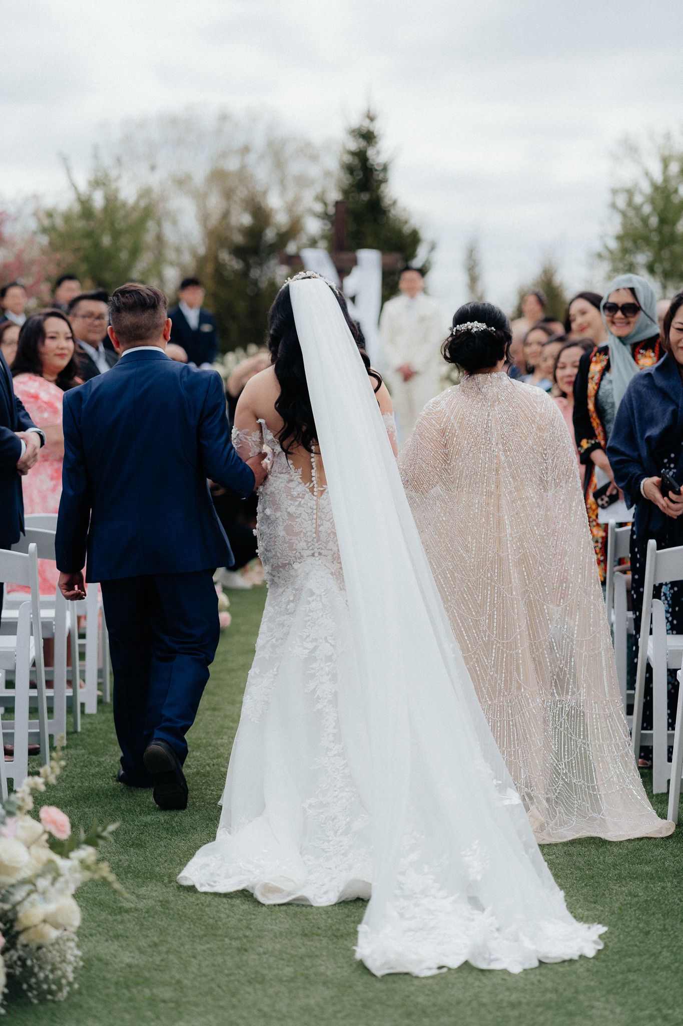 Wedding ceremony on a garden lawn with white chairs and guests watching as couple walks down the aisle.