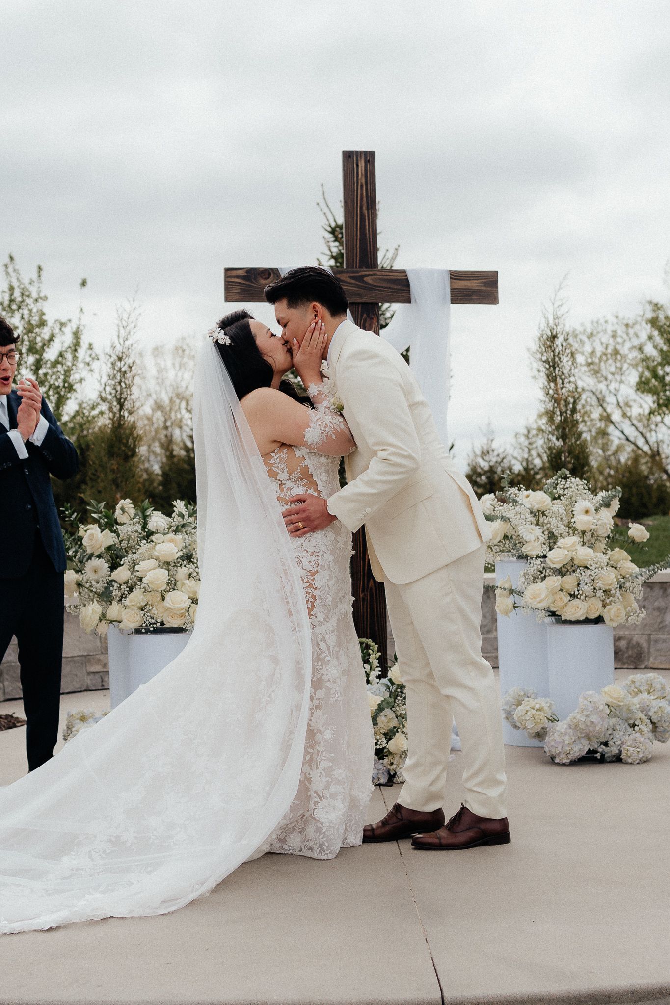 A couple shares their first kiss as newlyweds in front of a wooden cross with white floral decorations.