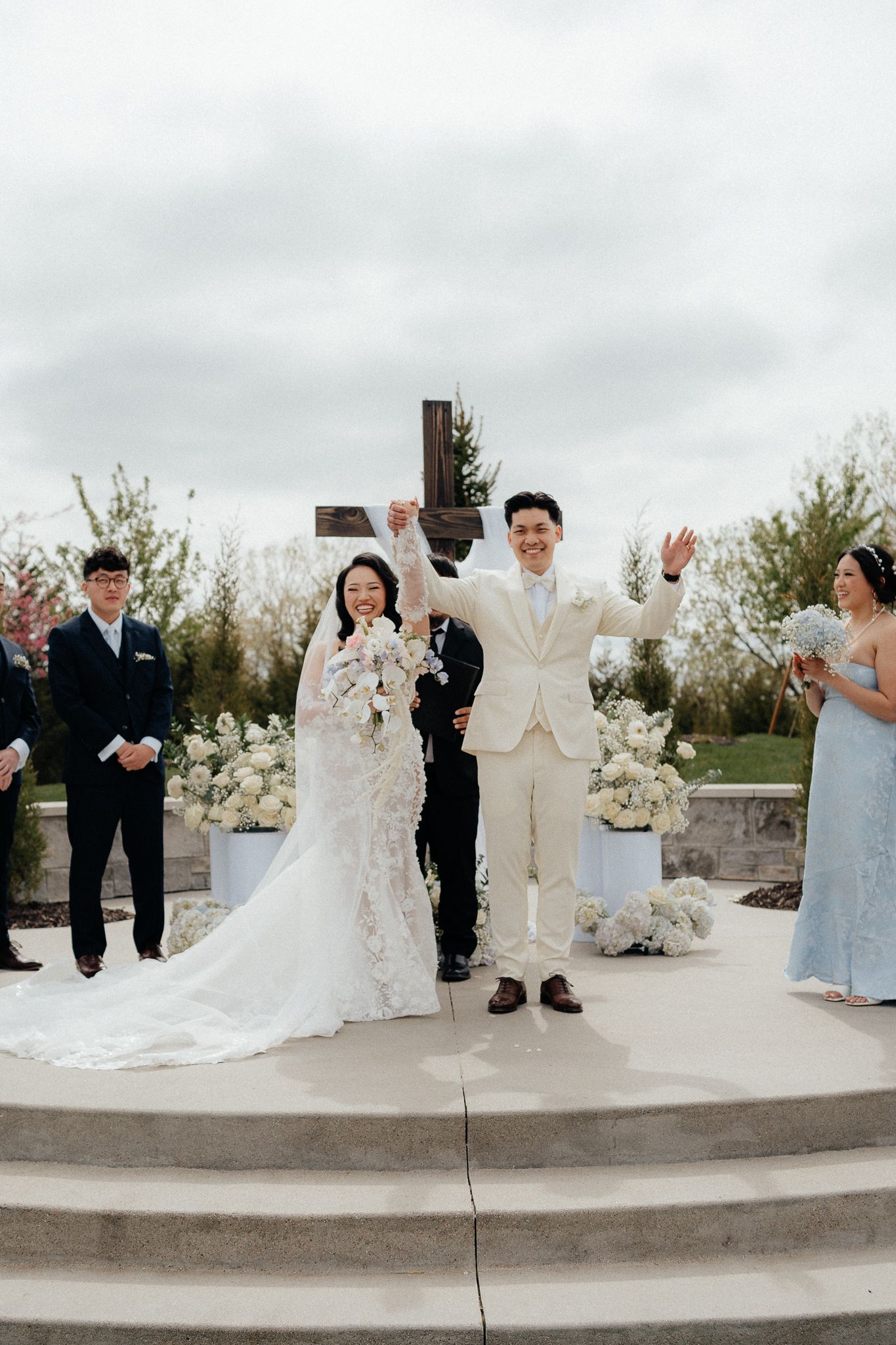 A couple celebrates at their outdoor wedding ceremony in front of flowers and a wooden cross.