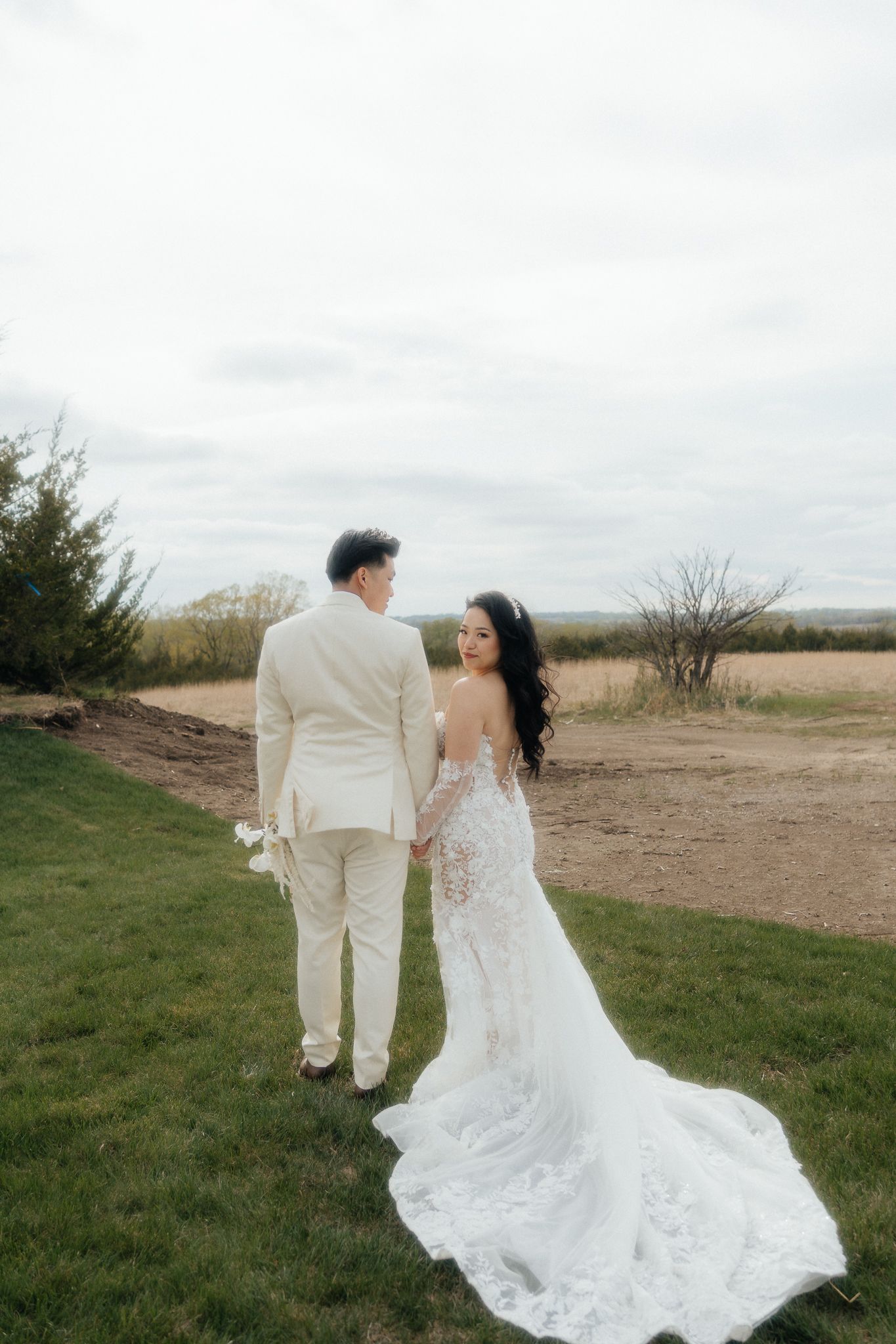 A couple holds hands in an outdoor wedding photo with a stunning lace wedding dress trailing on grass.