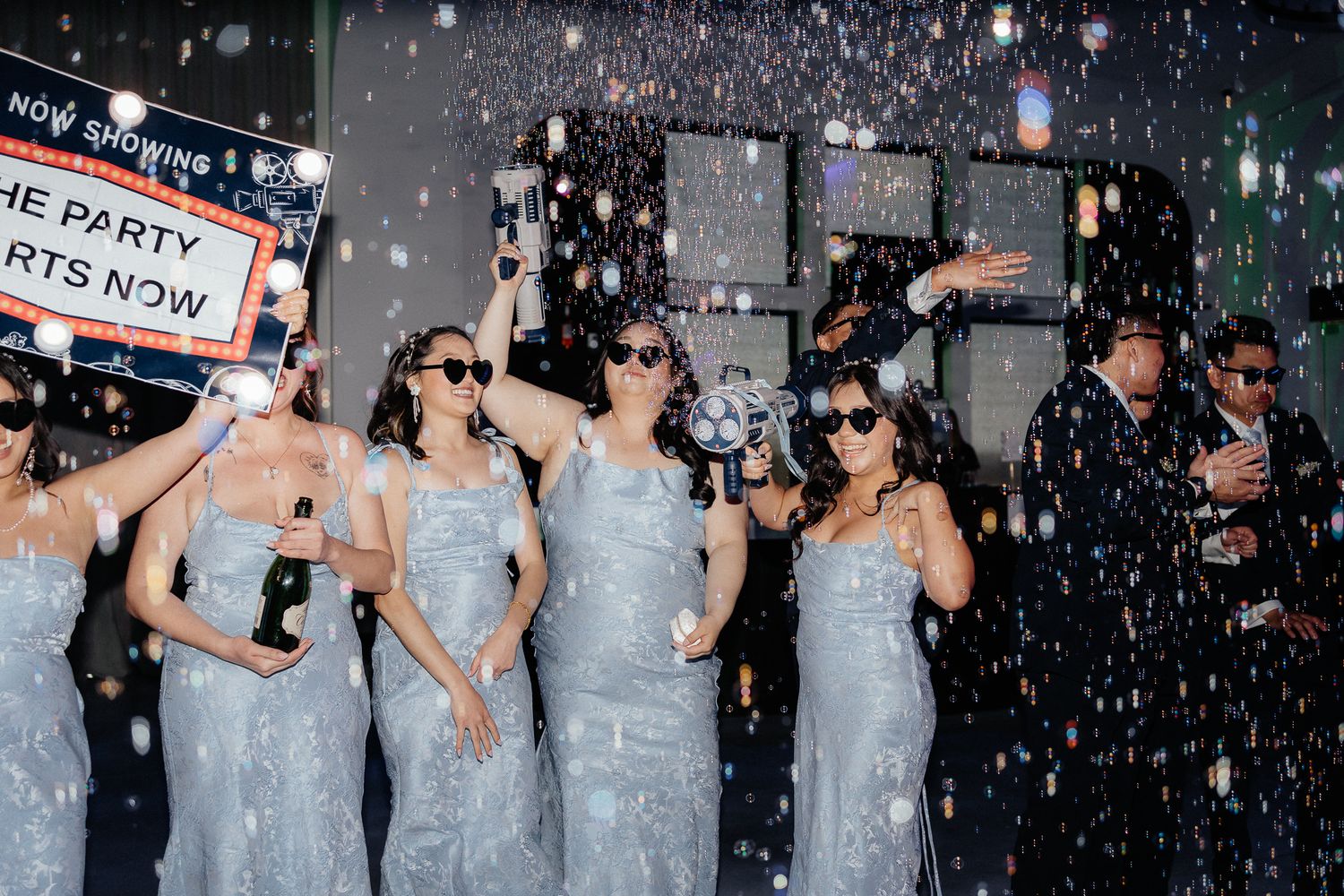 Bridesmaids in light blue dresses celebrate under falling confetti at a wedding reception.