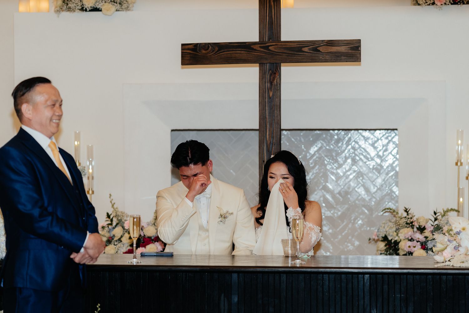 Emotional wedding reception moment at head table with couple and officiant standing beneath wooden cross.