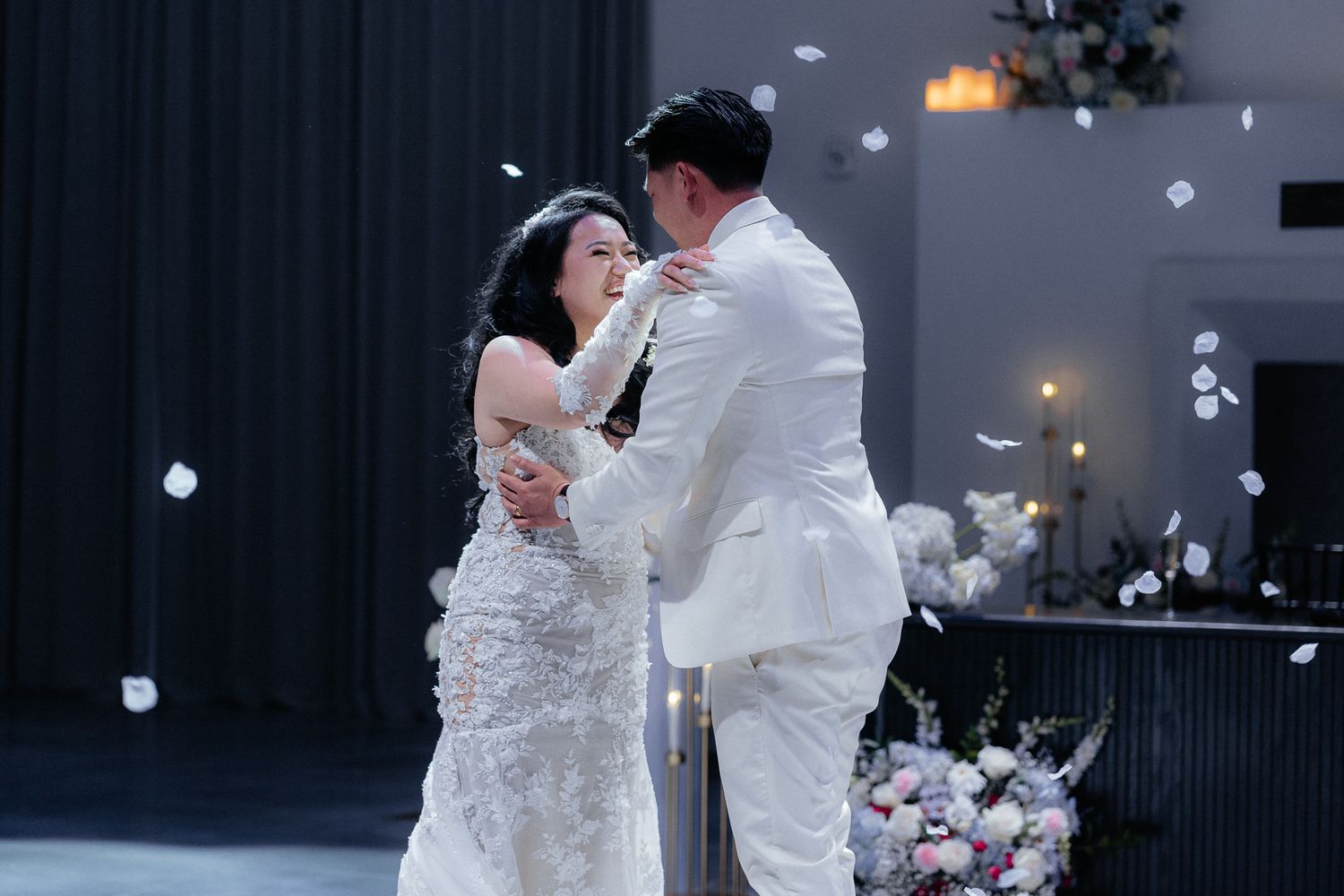 A couple in elegant wedding attire embrace during their romantic first dance surrounded by floating petals.