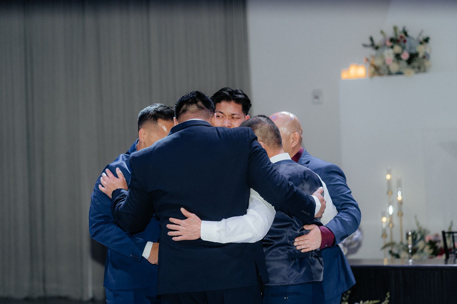 A group of men in navy blue suits embrace in a emotional circle hug during an indoor event.