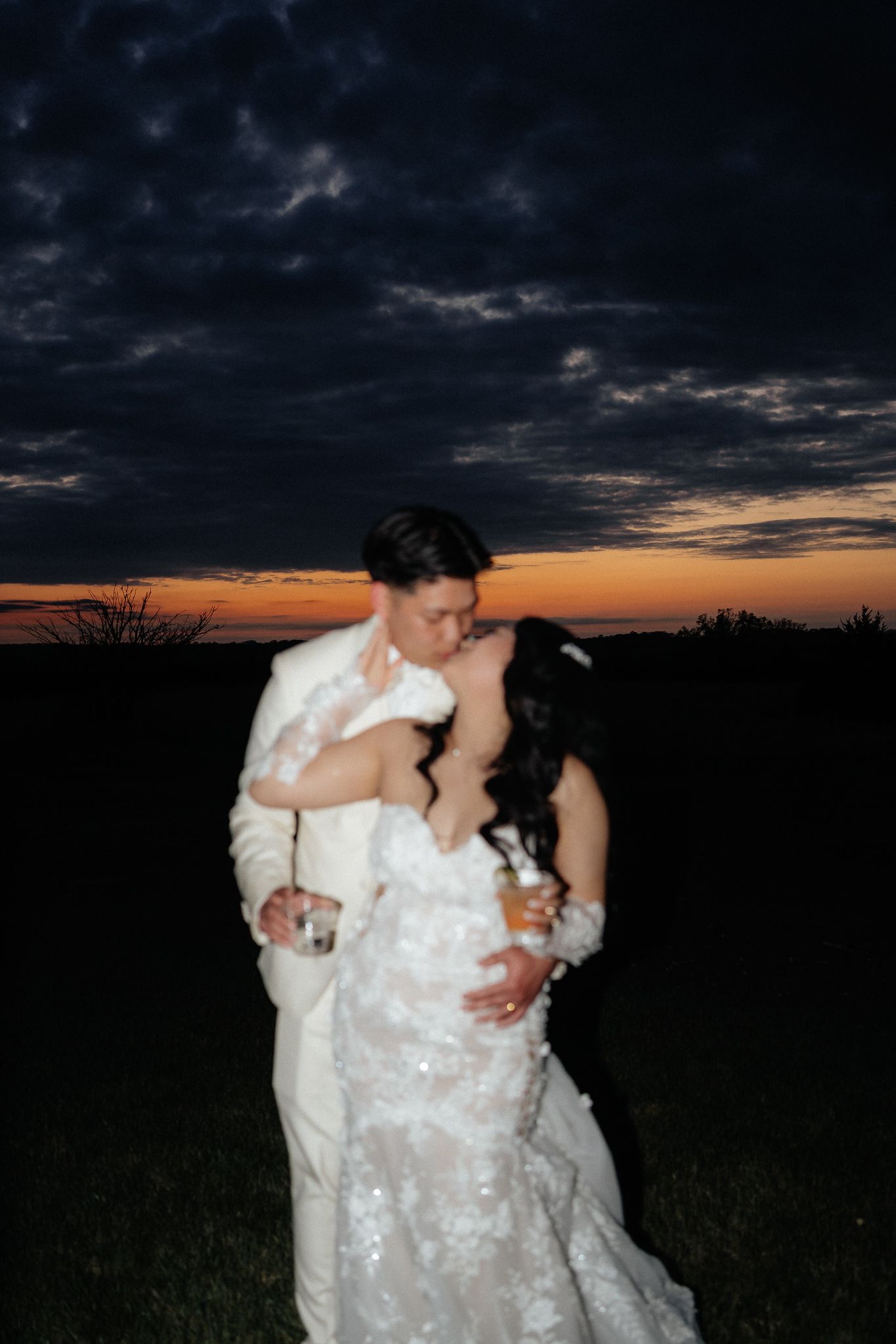 A couple embraces during sunset against a dramatic dark sky with orange horizon in their wedding attire.