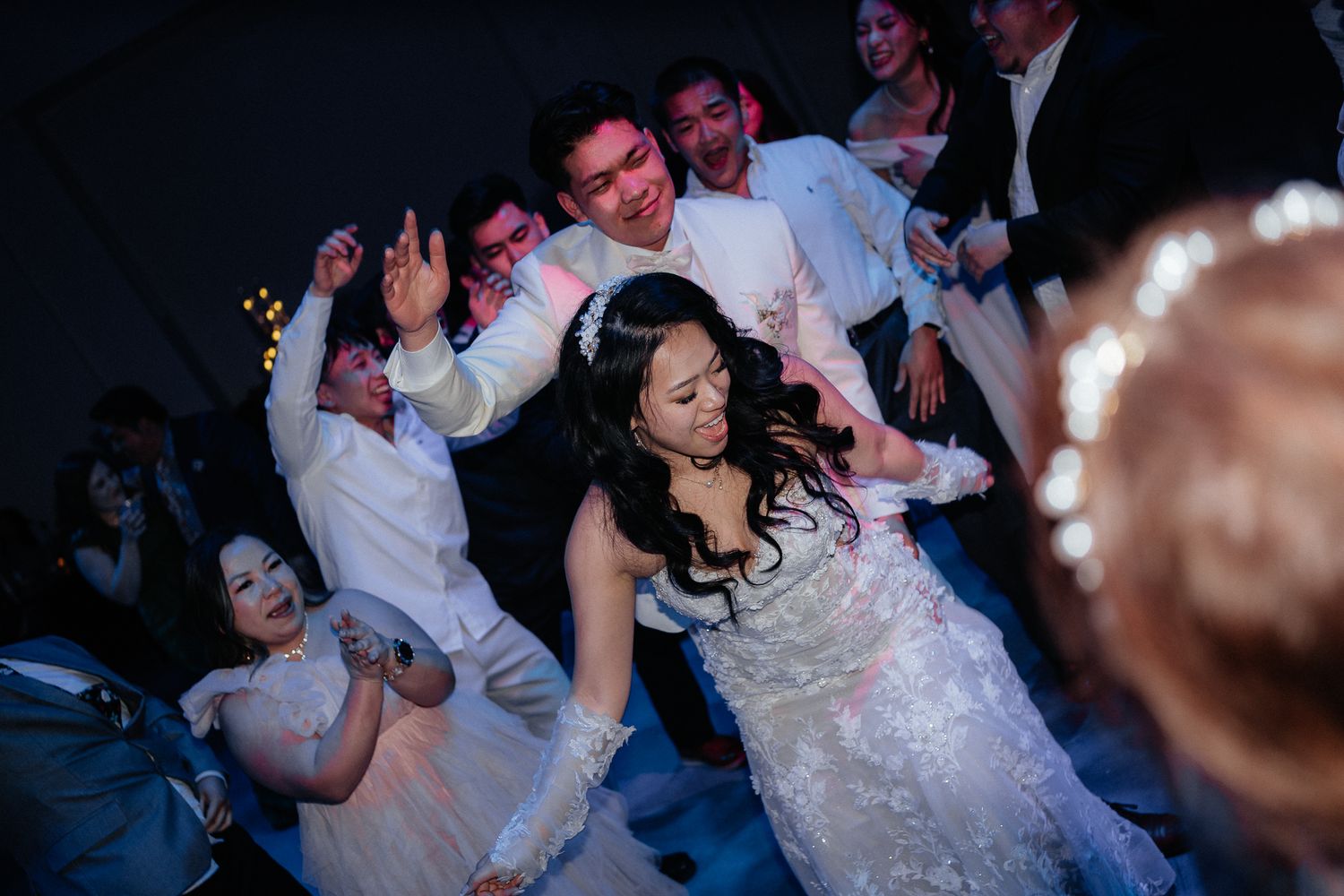 Wedding celebration on dance floor with guests in white attire dancing energetically around a bride in a flowing gown.