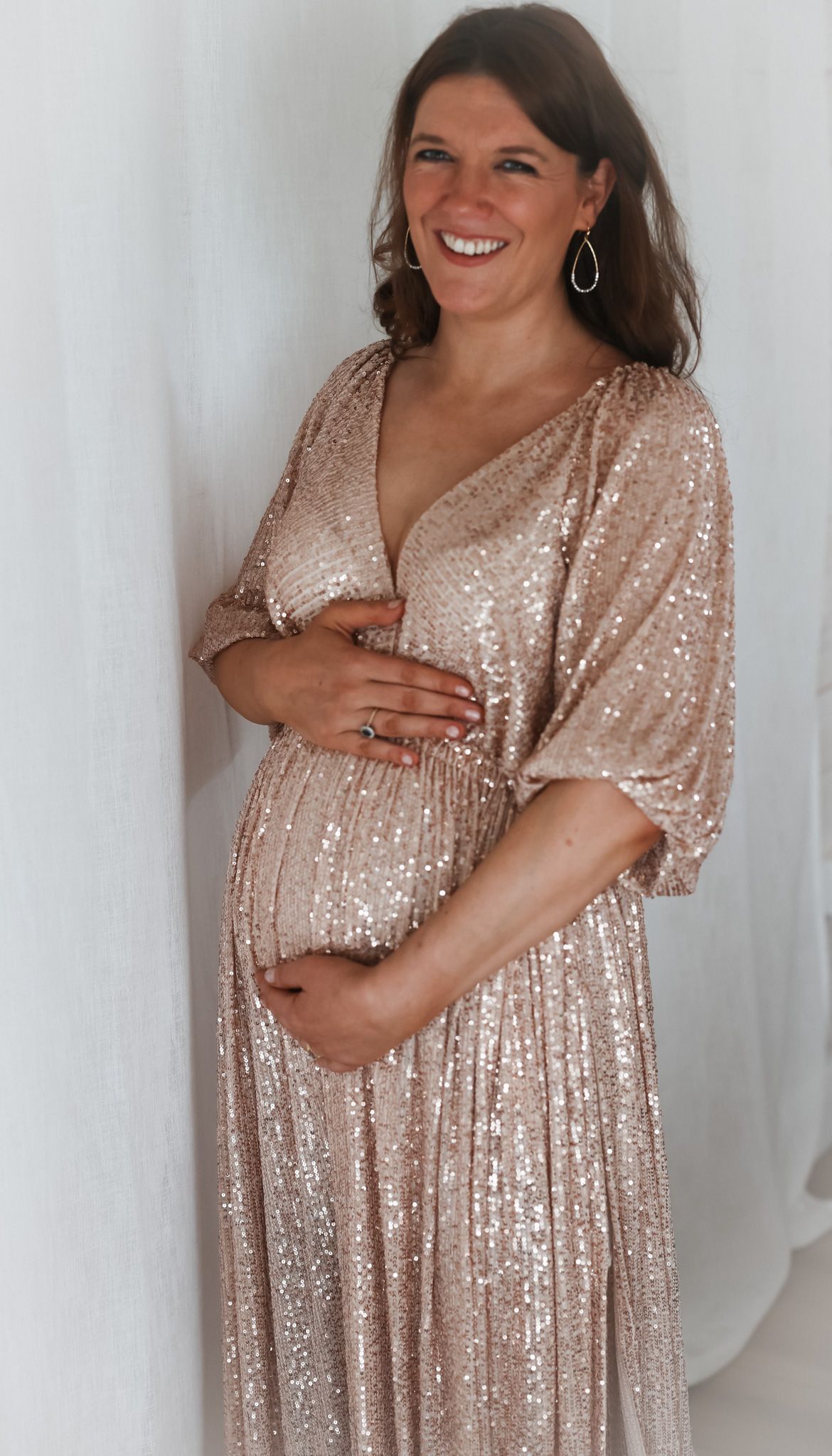 Pregnant woman in sparkly rose gold maternity dress touching belly against white wall.