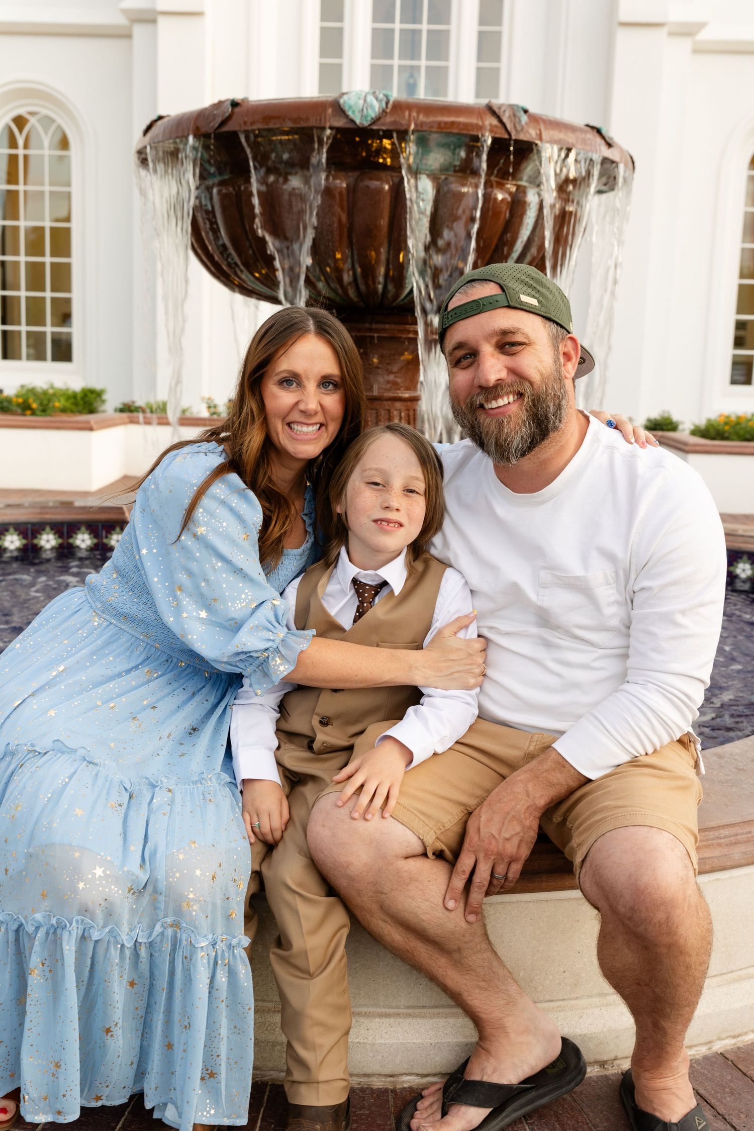 mom and dad sitting on either side of a boy sitting on edge of water fountain. wearing Sunday best