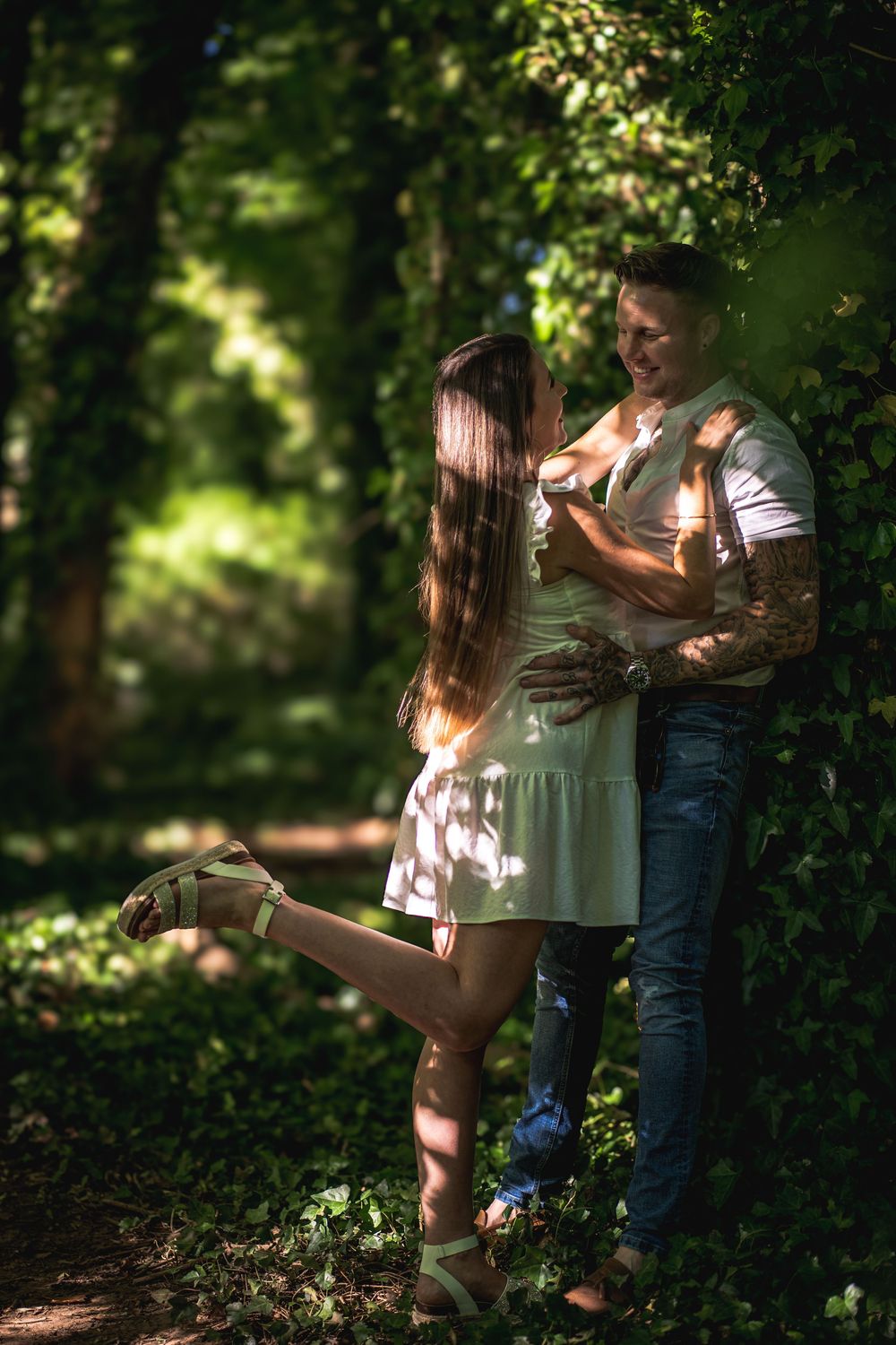 Couple in white outfits embracing in sunlit forest setting.